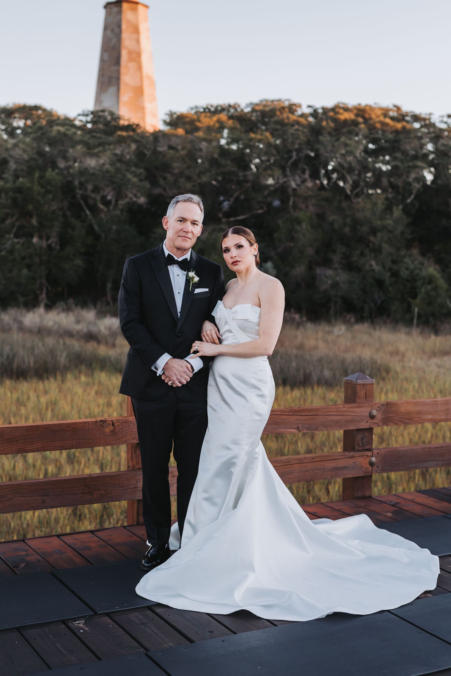 Bride in white mermaid gown and groom in black tuxedo pose on wooden deck with lighthouse backdrop at sunset.
