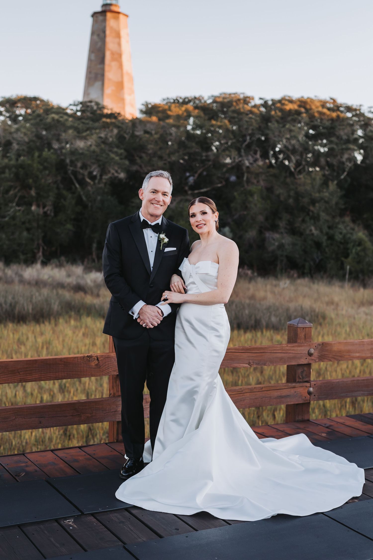 Bride and groom pose together on a wooden deck with a historic lighthouse and golden meadow backdrop at sunset.