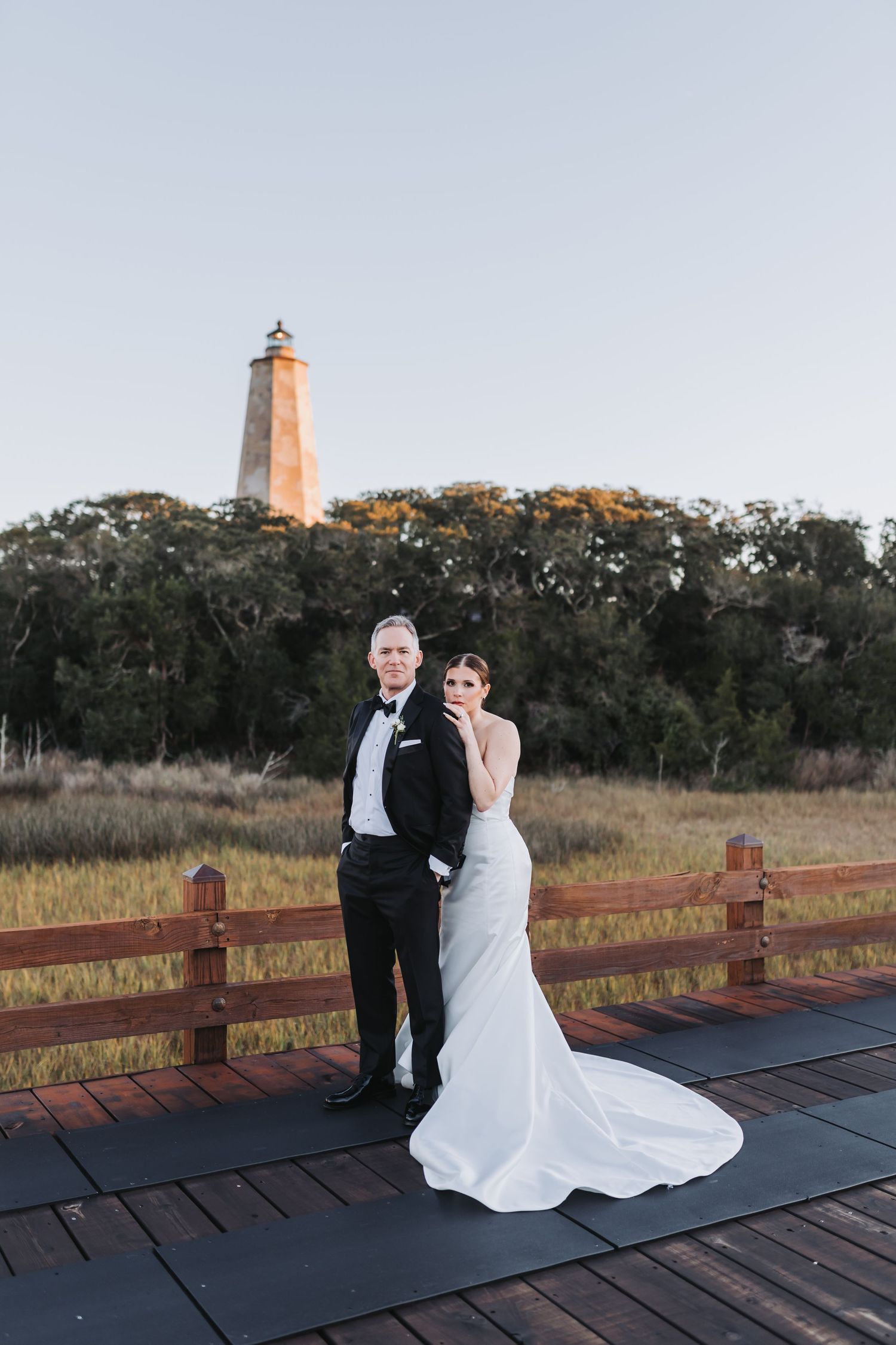 Bride and groom stand together on boardwalk with tall lighthouse and natural landscape at dusk.