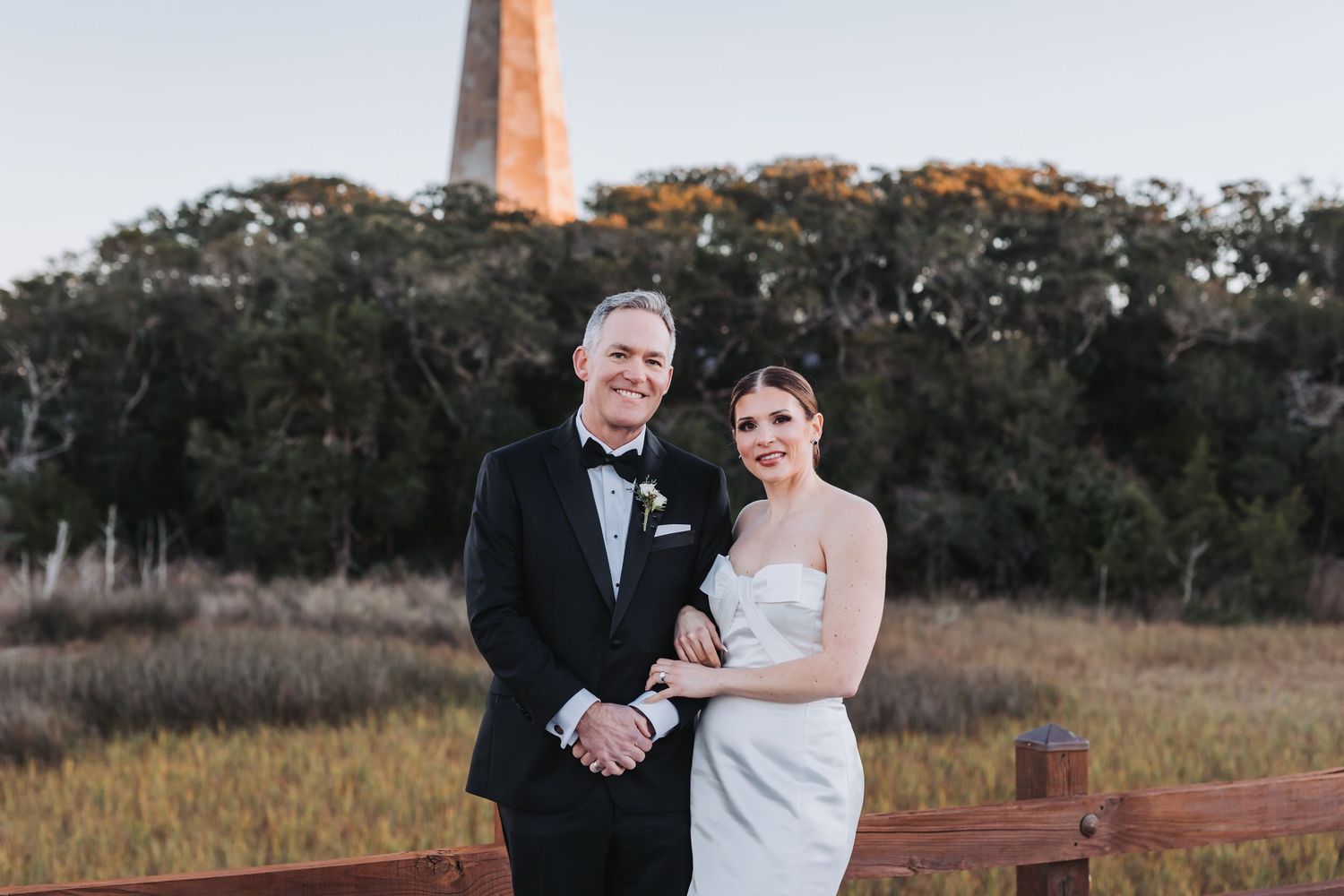A bride and groom smile together on a wooden fence with a lighthouse and marsh in the background.