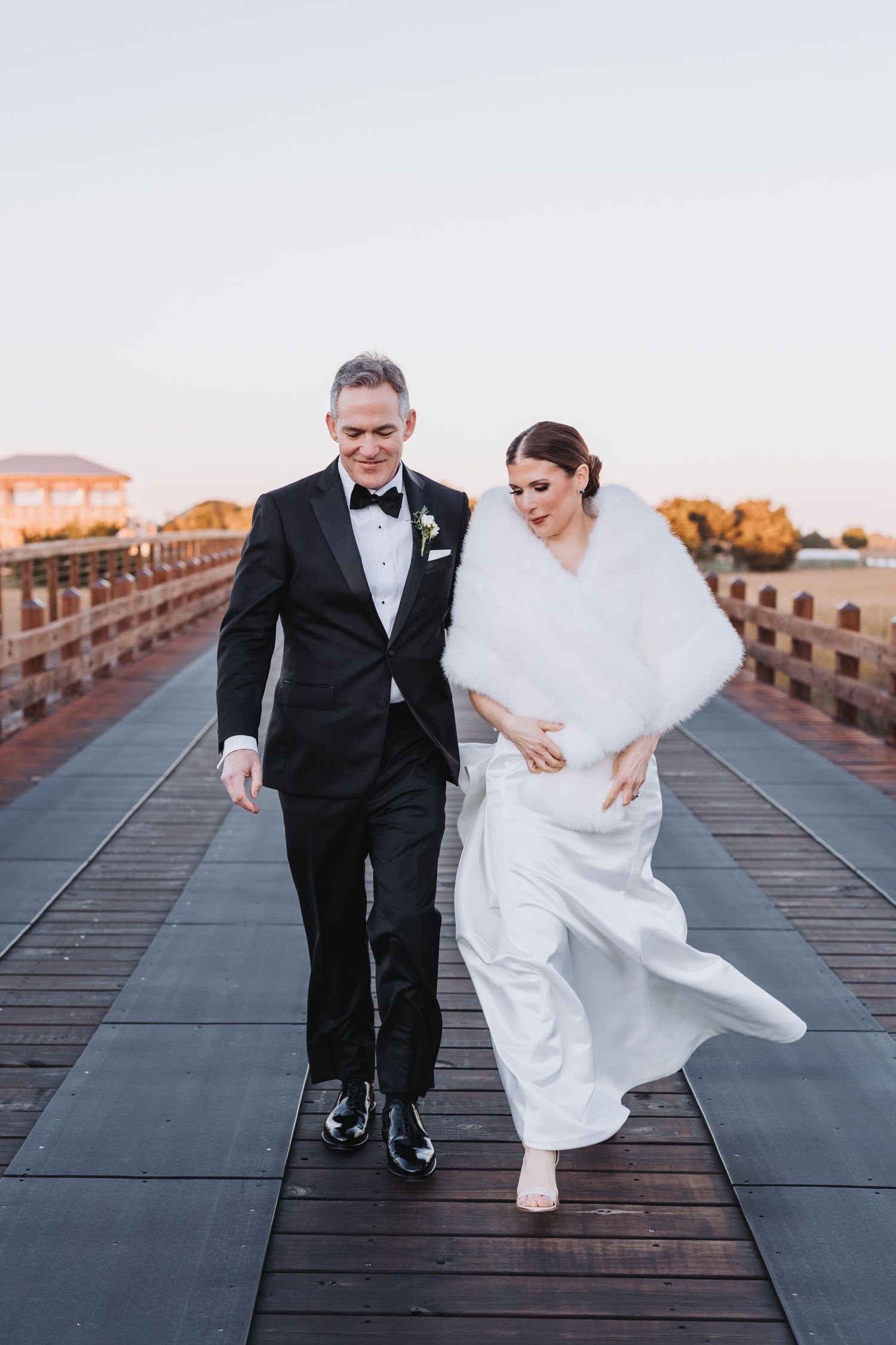 Bride and groom walking and laughing together on a waterfront boardwalk at sunset after their wedding.