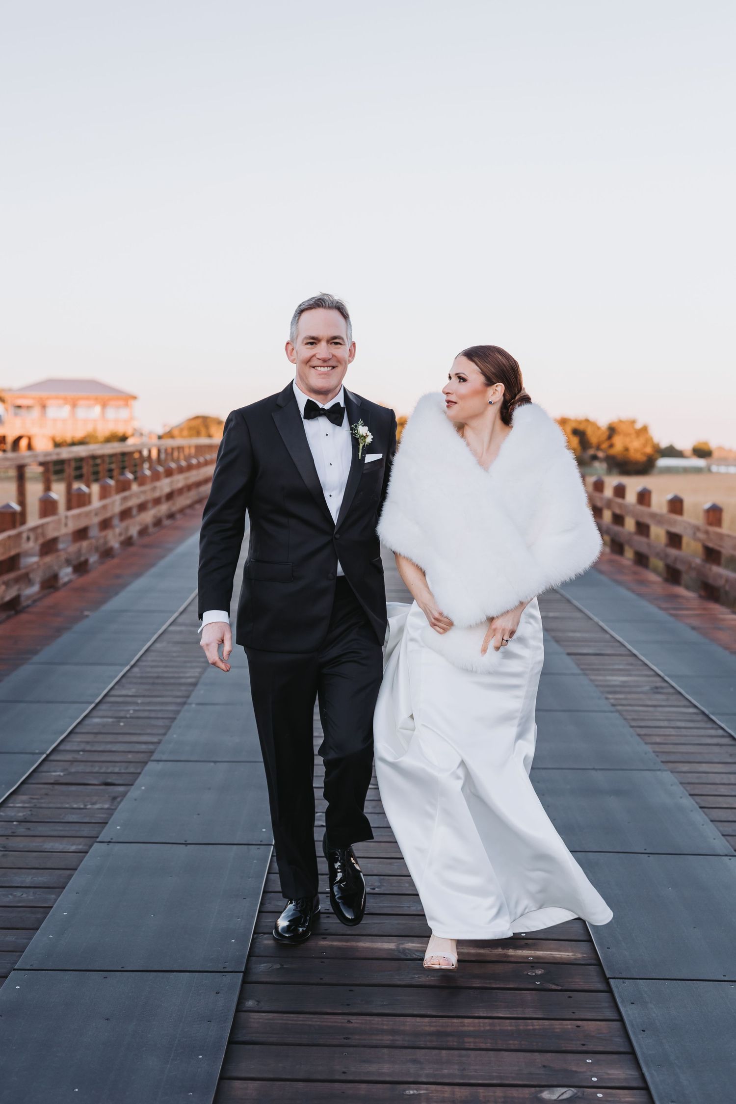 Bride and groom walking hand in hand on a waterfront pier at sunset, bride wearing white gown with fur wrap.