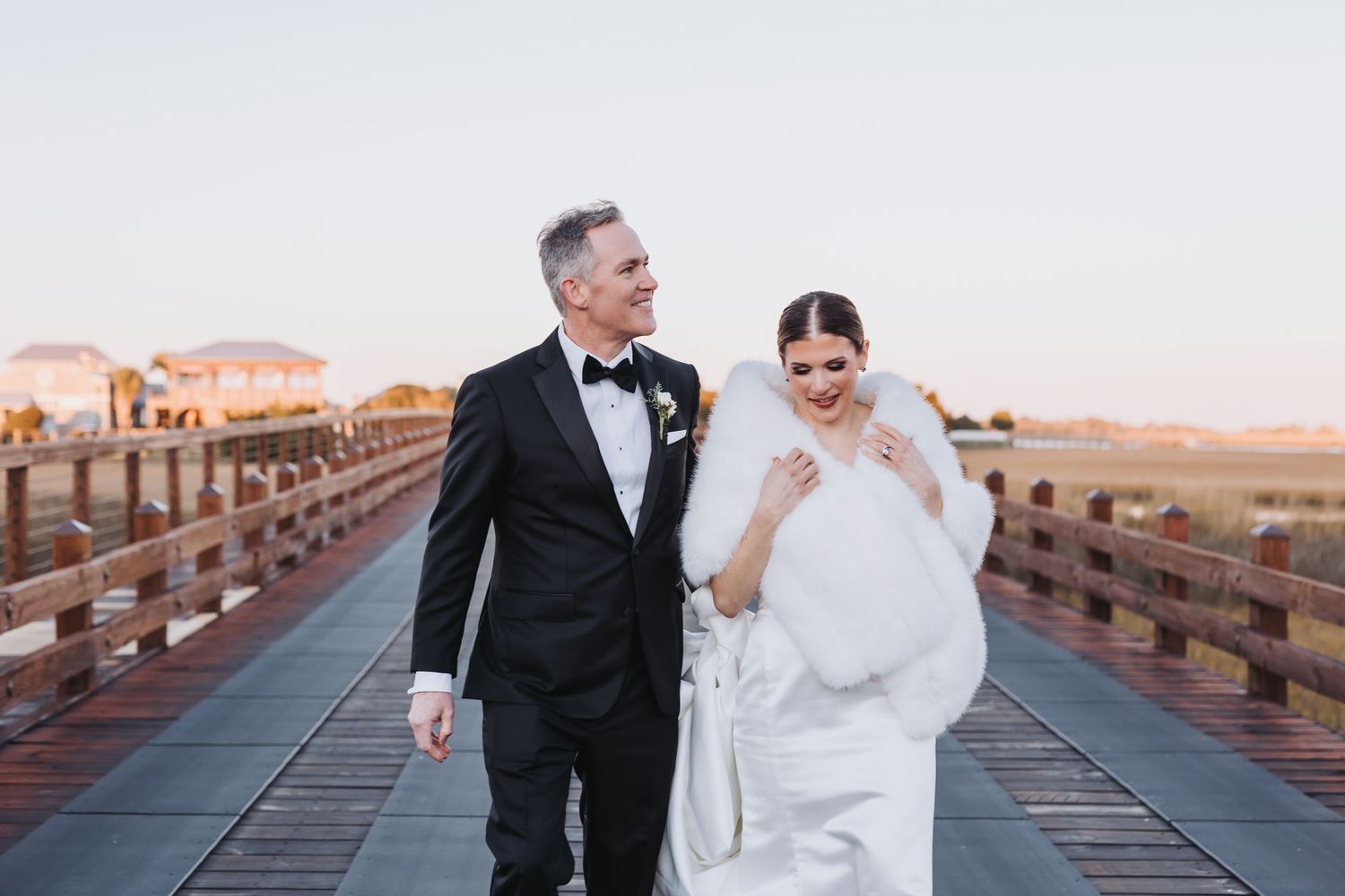 Bride in white gown and fur wrap walks with groom in tuxedo on wooden bridge at sunset.