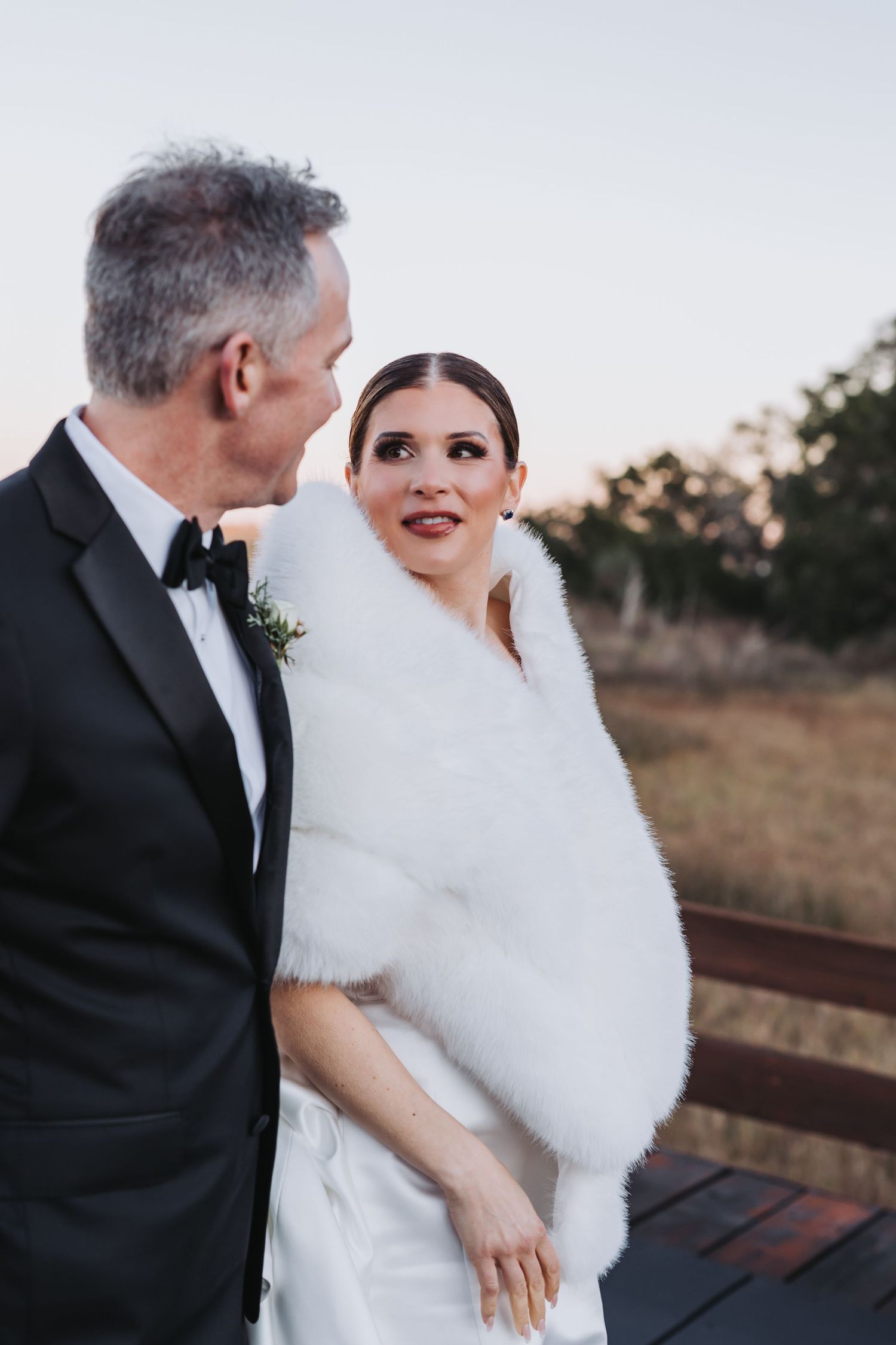 Bride in white fur stole and groom in tuxedo share a romantic moment outdoors at their winter wedding.