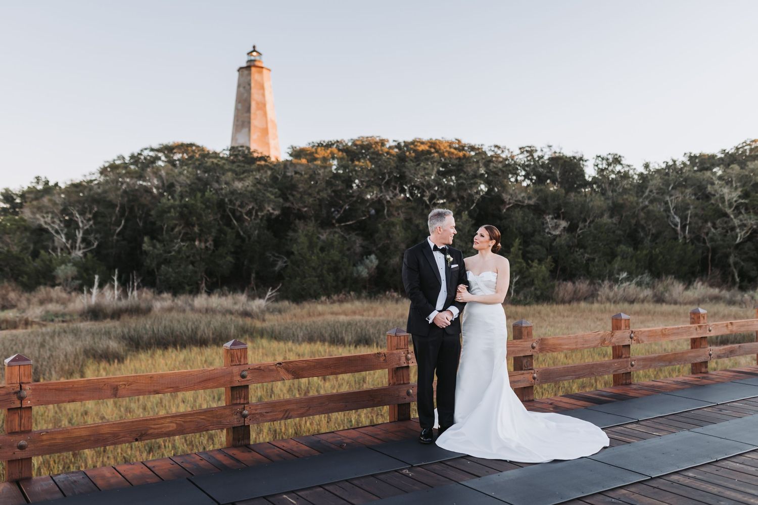 Bride and groom pose on wooden boardwalk with historic lighthouse and golden marshland in background at sunset.