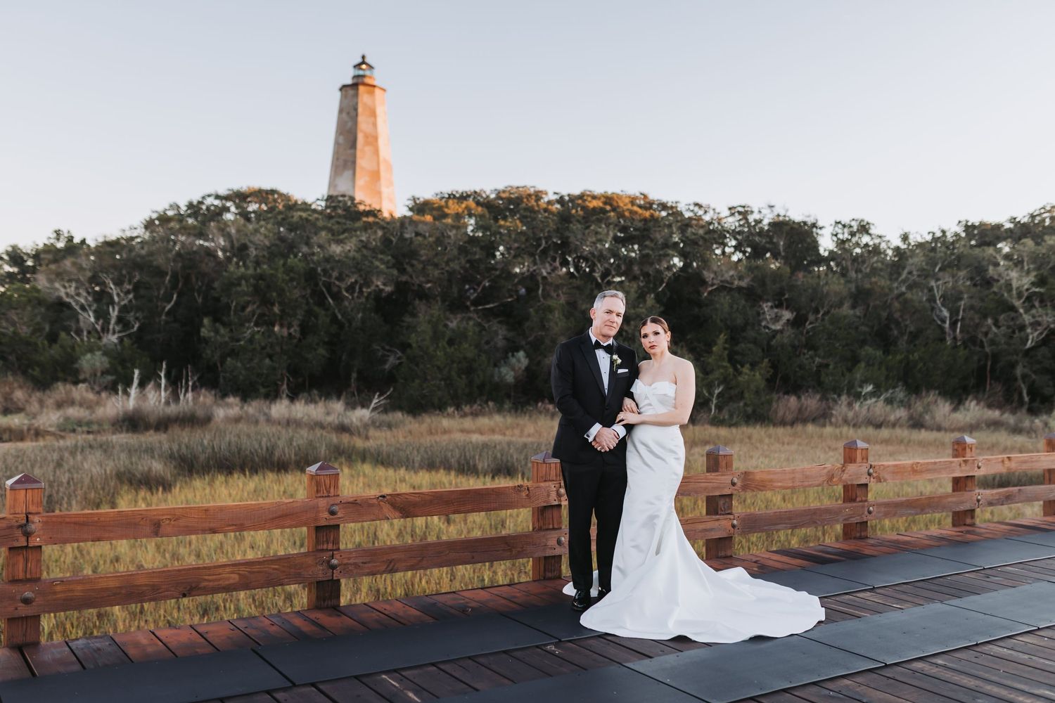 Bride and groom pose on wooden deck with historic lighthouse and autumn trees at golden hour outdoor wedding.
