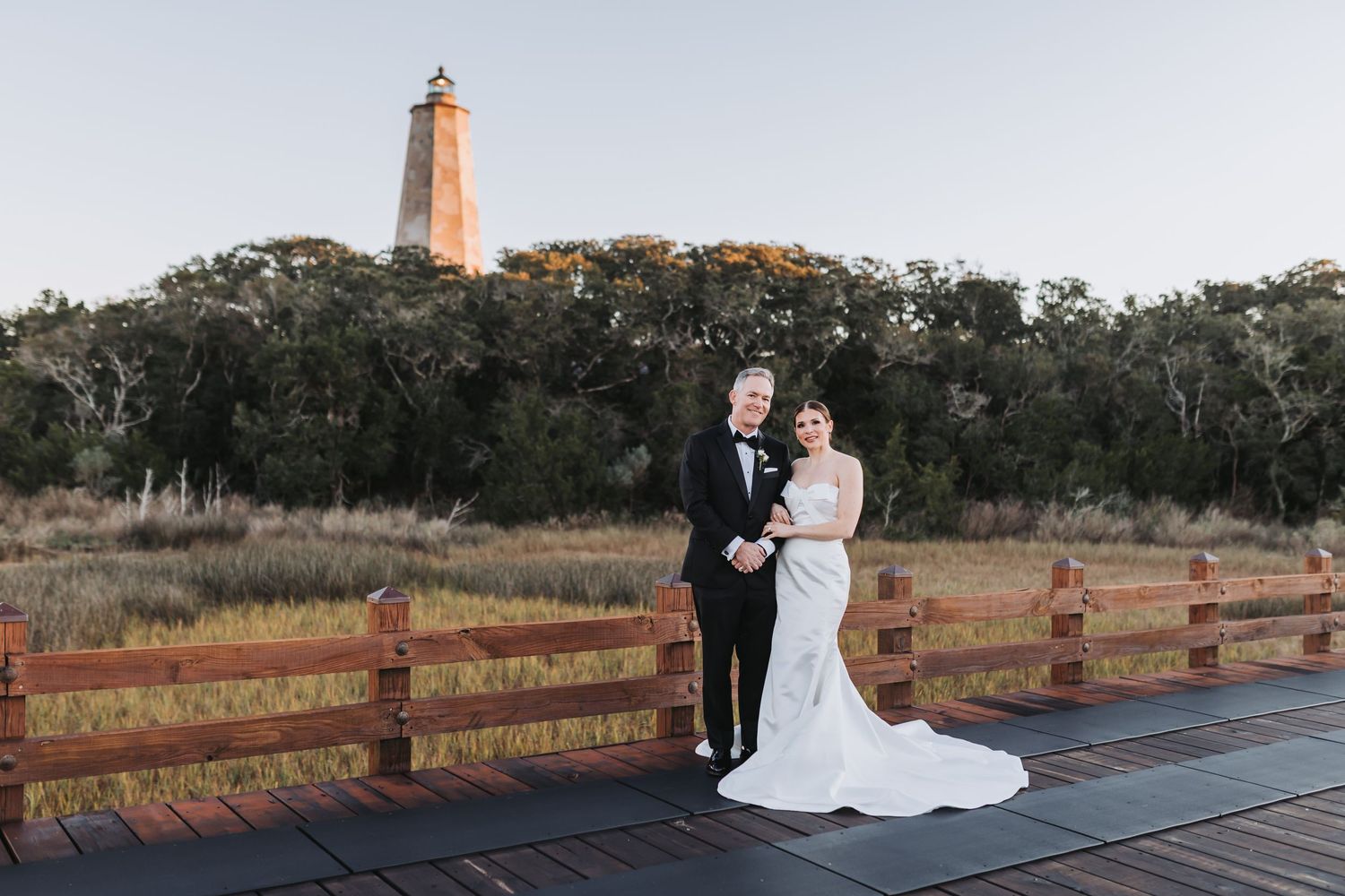 Bride and groom pose on wooden deck with historic lighthouse and trees in background at golden hour.