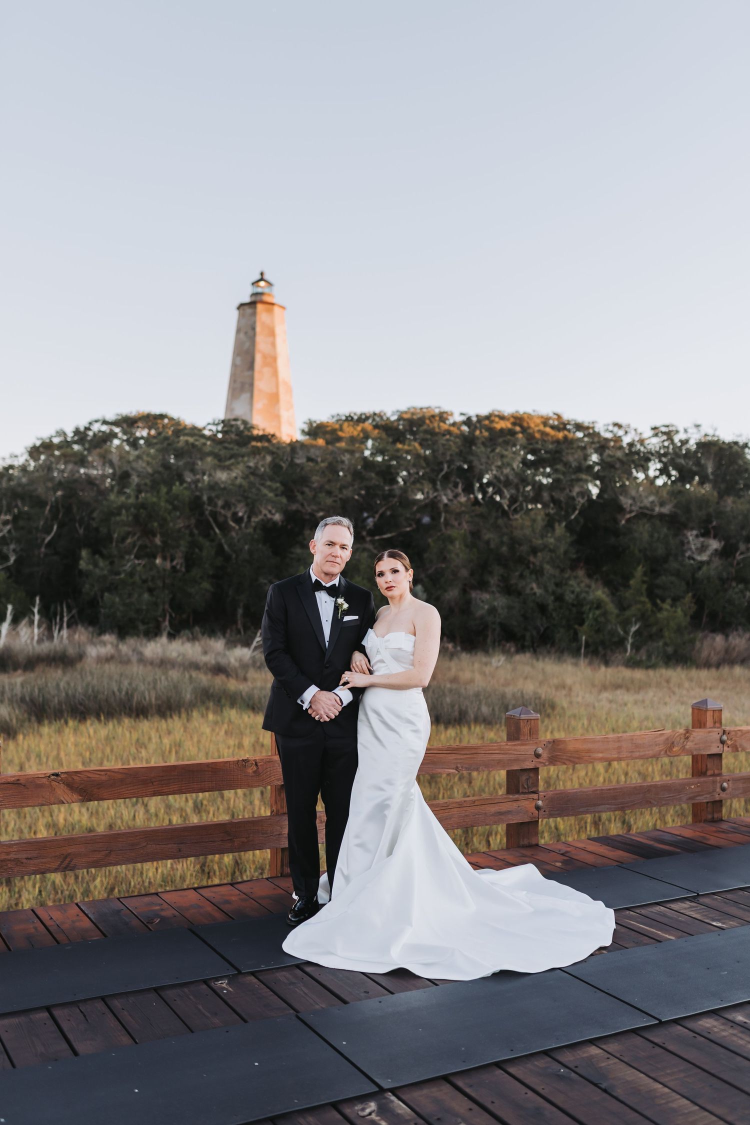 Bride and groom pose on wooden deck with lighthouse backdrop at sunset wedding.