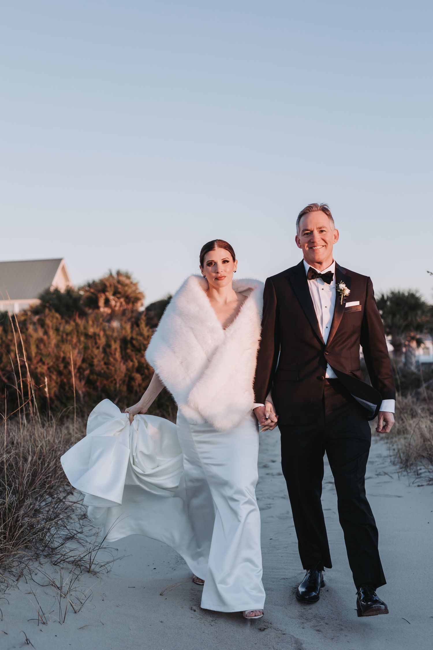 Bride in white jumpsuit with fur stole and groom in burgundy tuxedo walking hand in hand on a sandy beach at sunset.