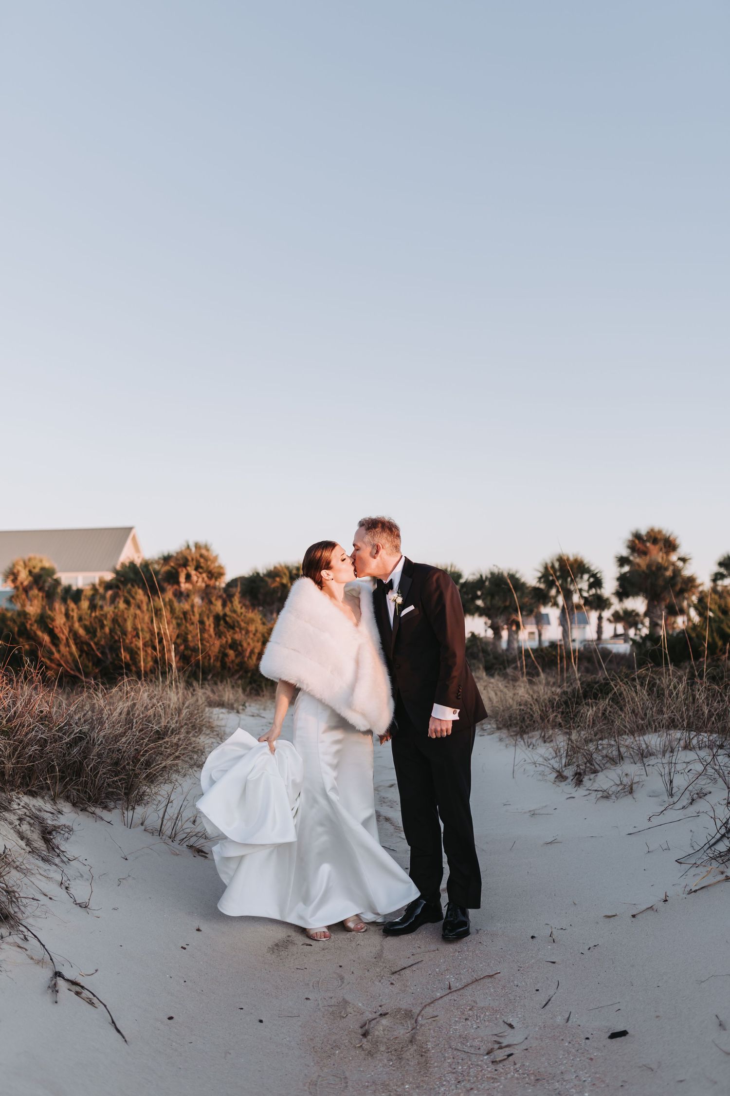 Bride and groom share a romantic kiss on sandy beach dunes at sunset, bride wearing white gown with fur wrap.