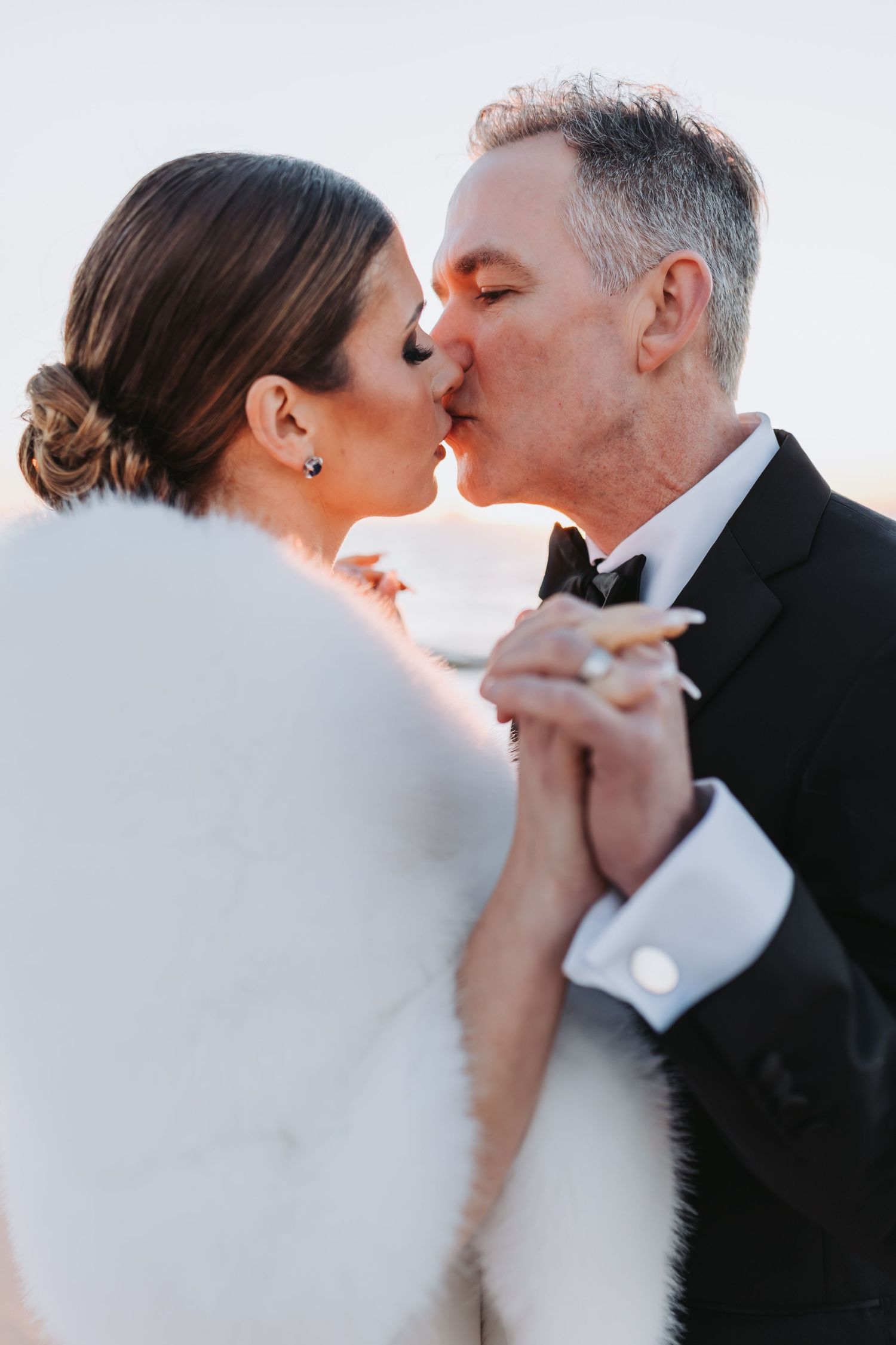 Bride in white fur stole and groom in tuxedo share a kiss at beach wedding.