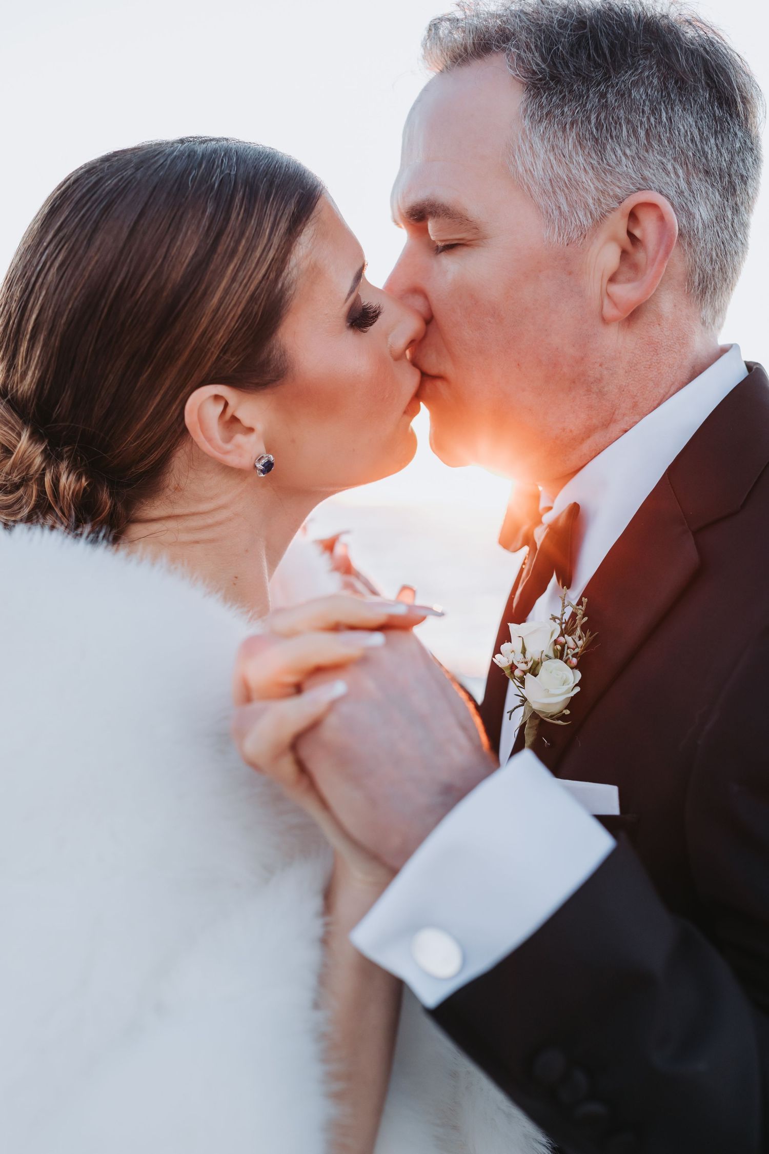 Bride and groom share a romantic sunset kiss on their wedding day, hands clasped together.