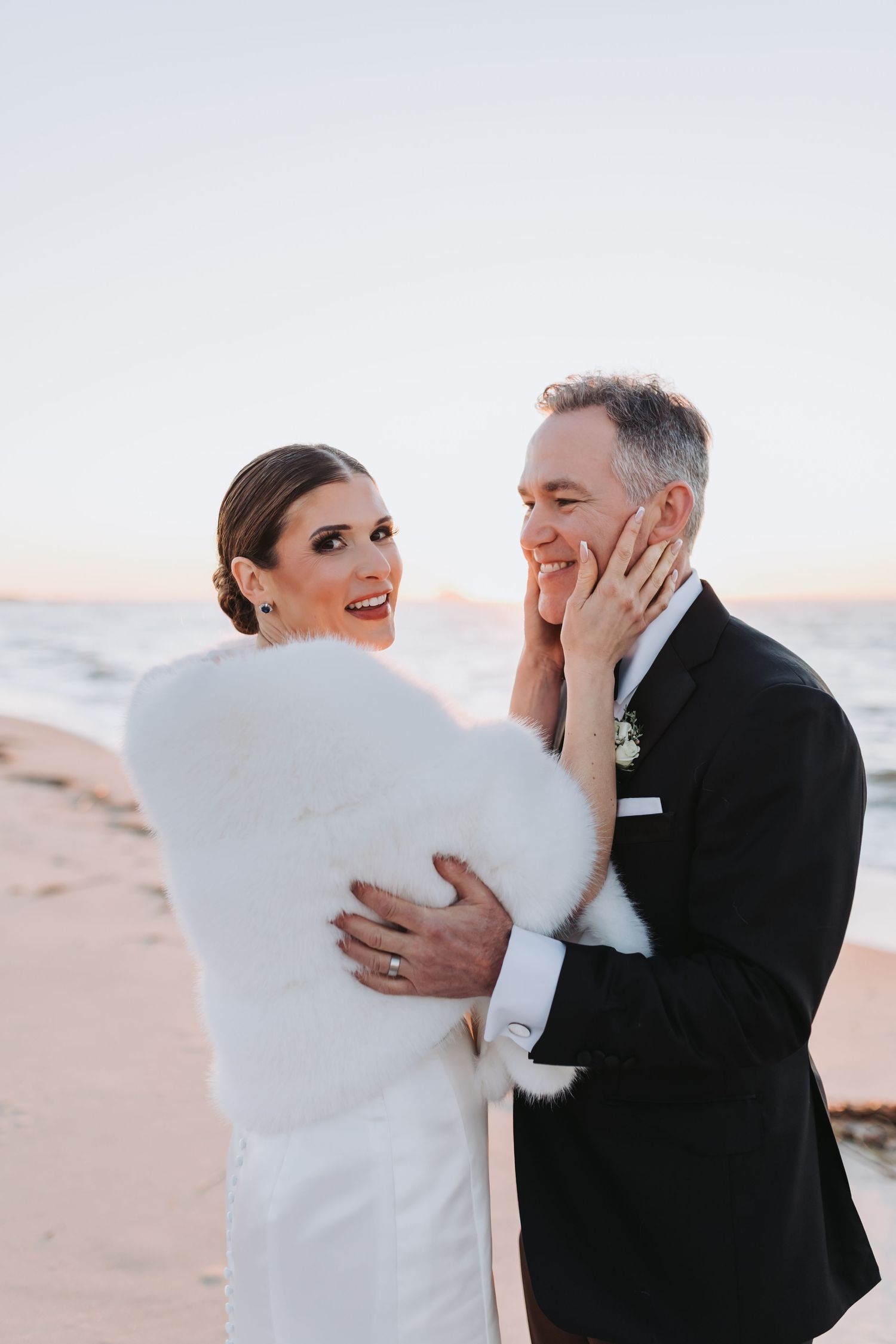 Bride in white fur stole laughs with groom in black tuxedo on sandy beach at sunset.