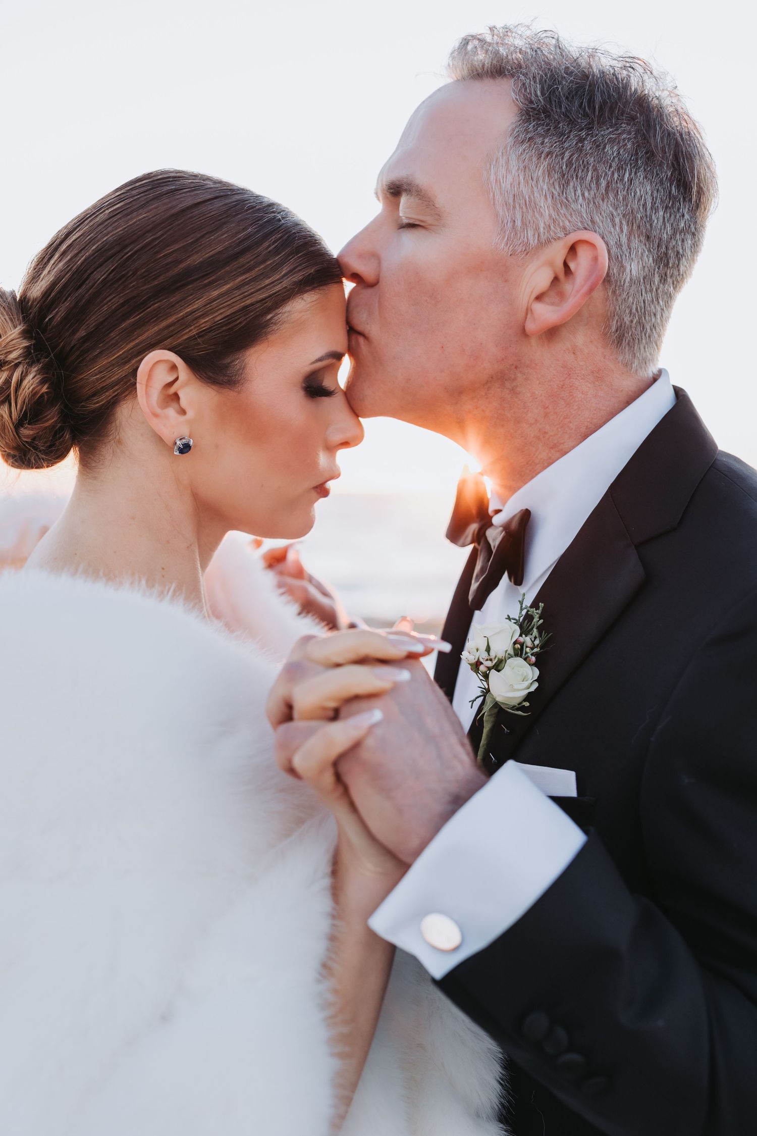 Groom tenderly kisses bride on forehead at sunset, bride wearing elegant fur stole and drop earrings.