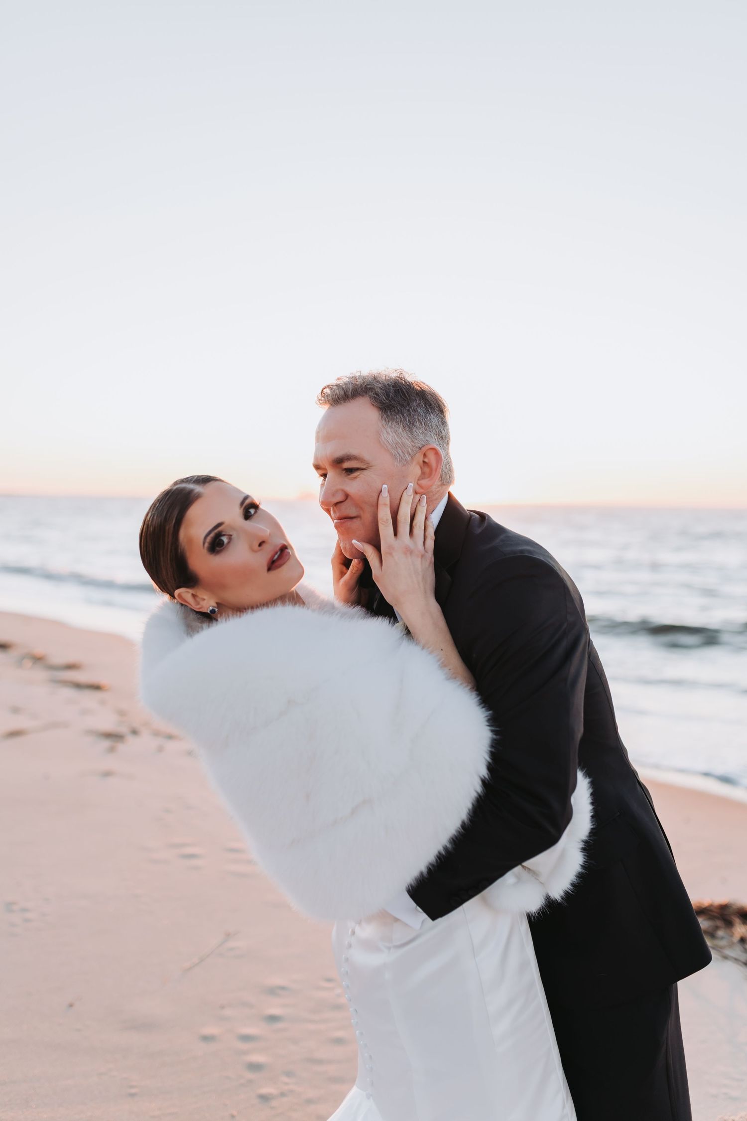 Bride in white fur stole embraces groom in black tuxedo on sandy beach at sunset.
