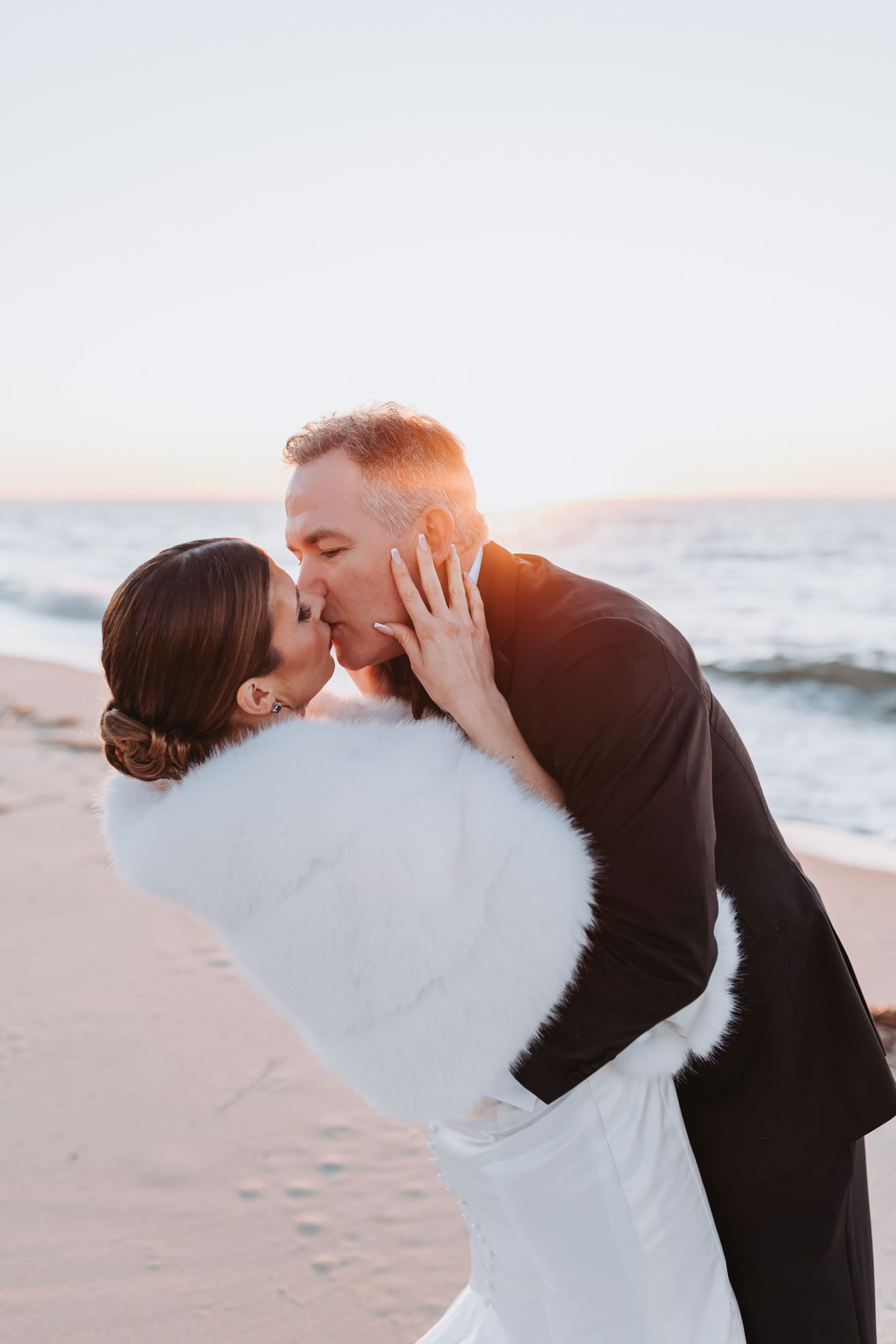 Bride and groom sharing a romantic kiss on the beach at golden hour, bride in white fur wrap.