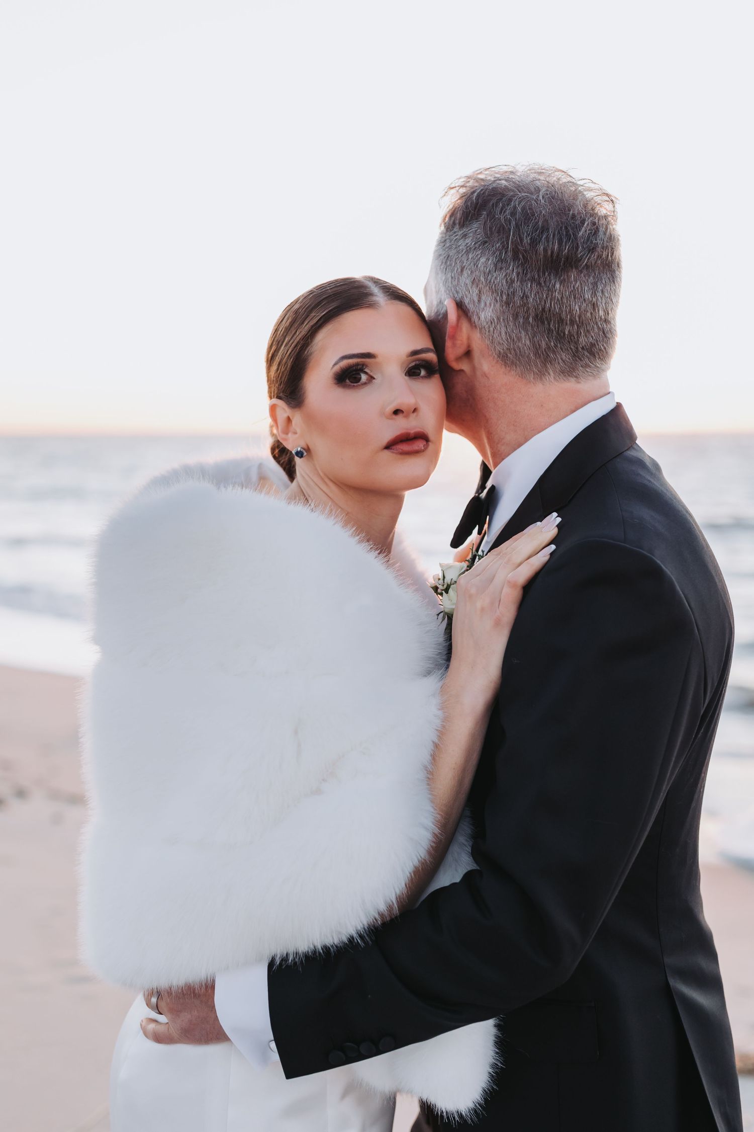 Elegant bride in white fur wrap embraced by groom on sandy beach at sunset.