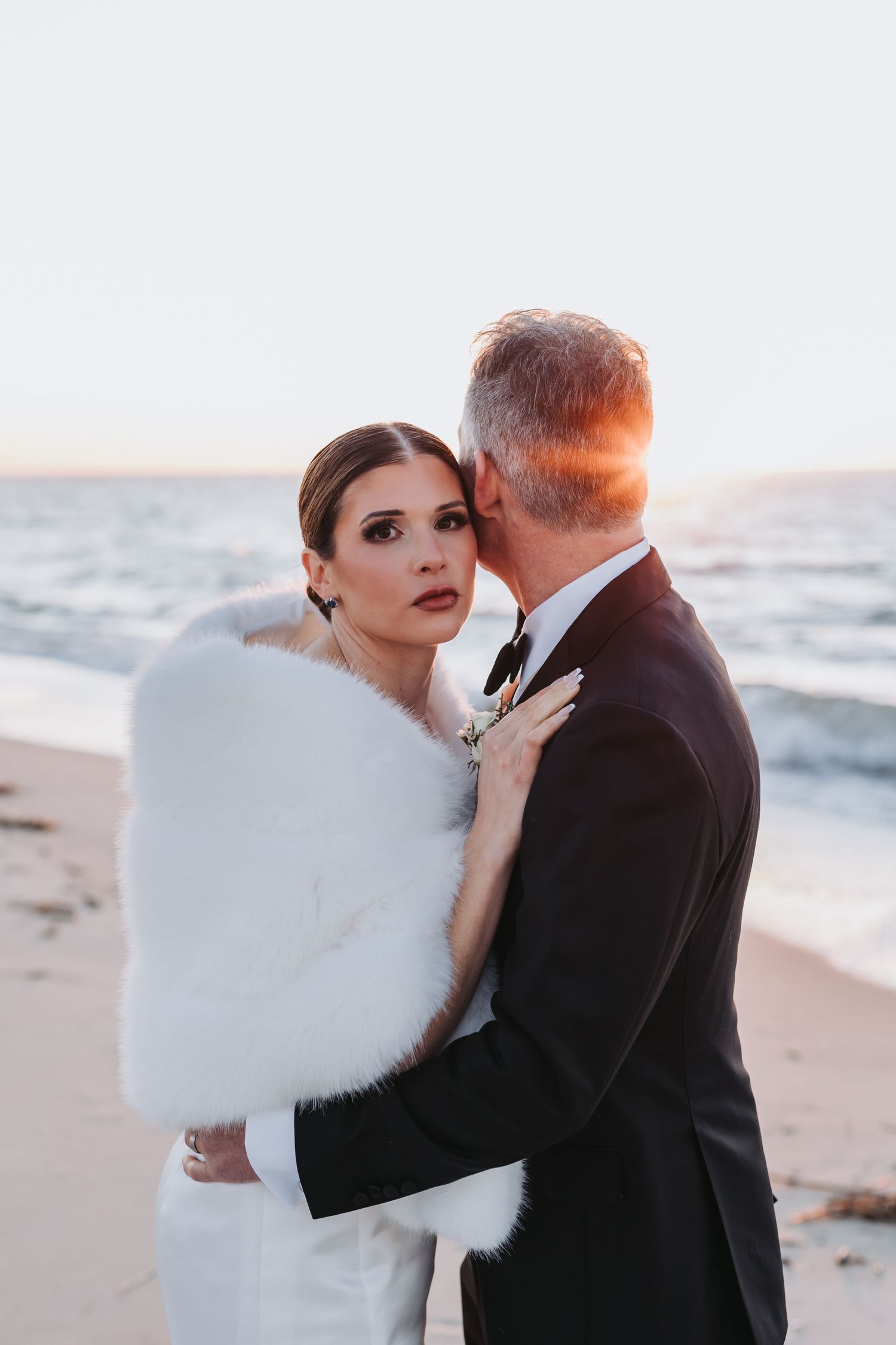 Groom kisses bride's cheek as she gazes at camera on beach during golden hour sunset.