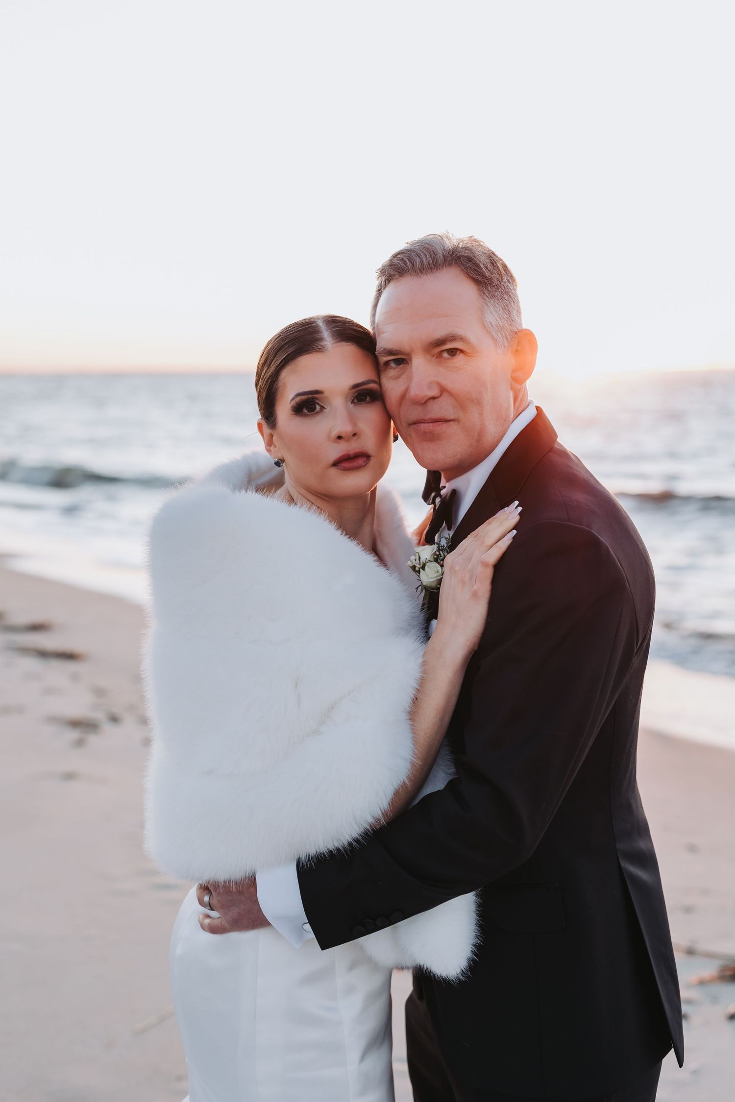 Elegant couple embraces on beach at golden hour, bride wearing white fur wrap and groom in tuxedo.