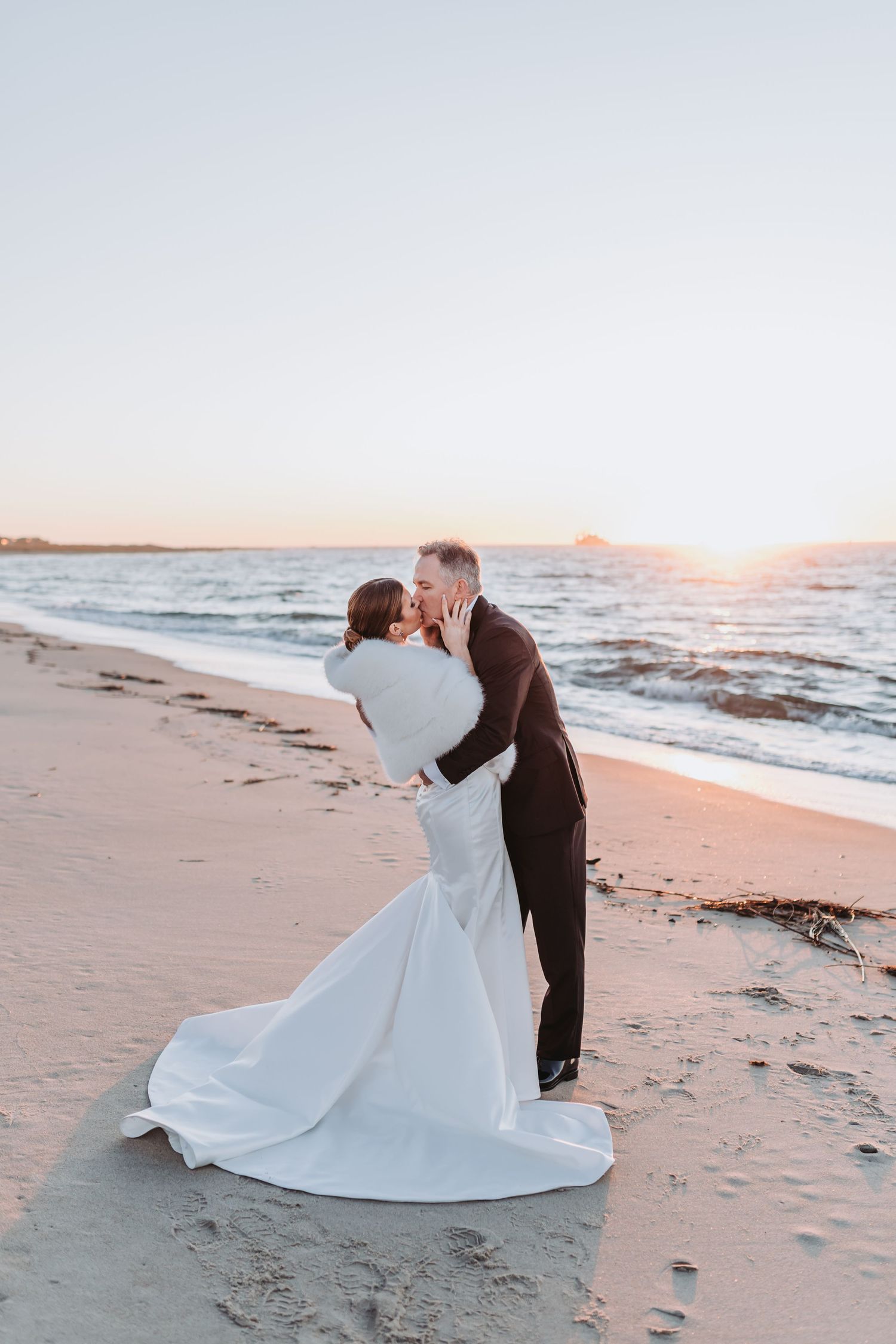 Bride and groom embrace in a romantic kiss on a beach at golden sunset, bridal gown train flowing on sand.