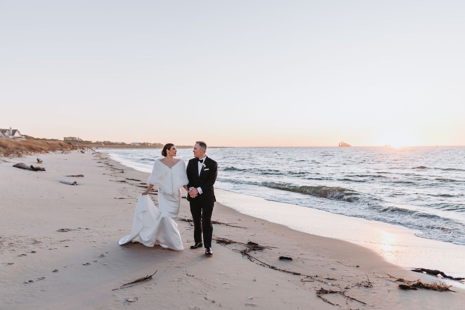 Bride and groom walk hand in hand along a sandy beach at sunset in wedding attire.