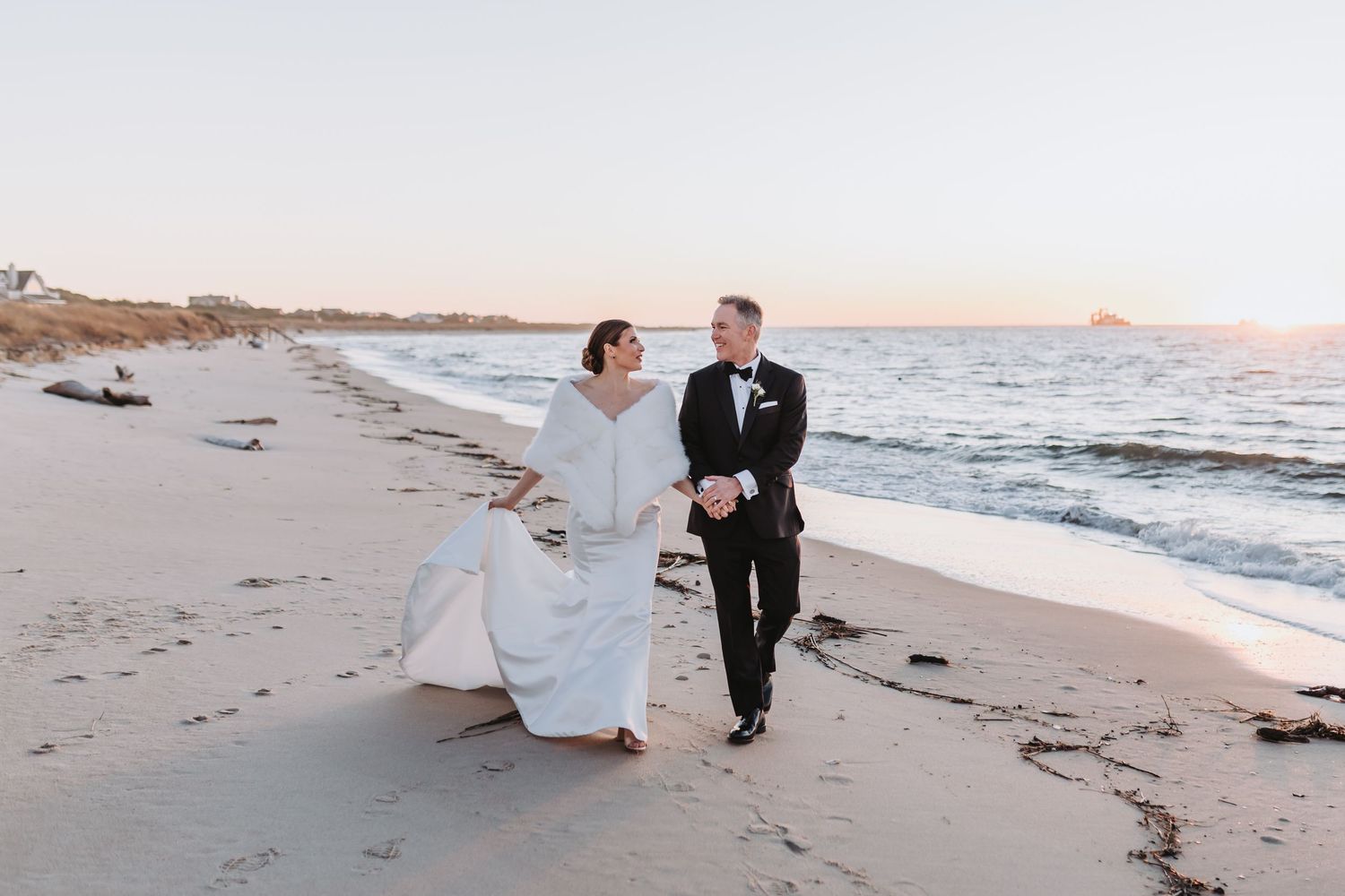 Bride and groom walk hand-in-hand along a sandy beach at sunset, dressed in wedding attire.