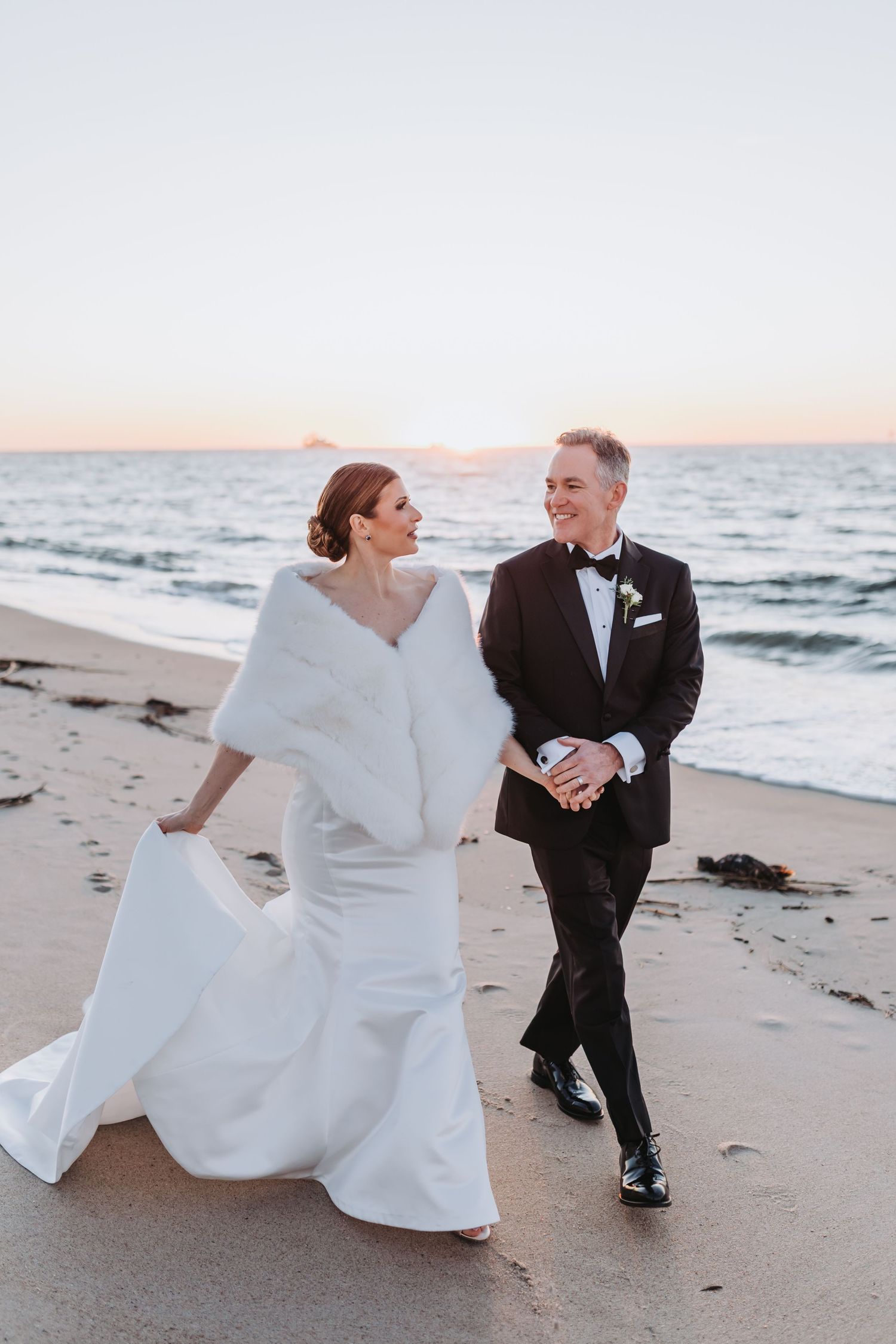 Bride in white gown and fur stole walks hand-in-hand with tuxedo-clad groom along sunset beach.