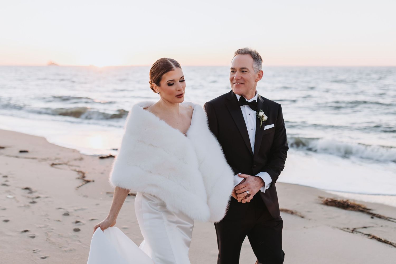 Bride in white gown with fur wrap and groom in tuxedo walking on beach at sunset.