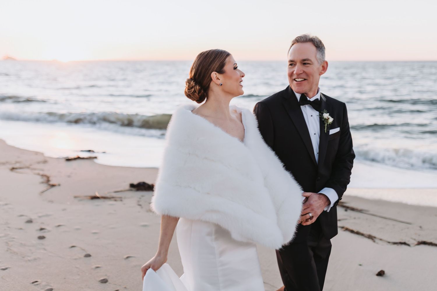 Bride in white gown with fur wrap and groom in tuxedo walking hand in hand on a beach at sunset.