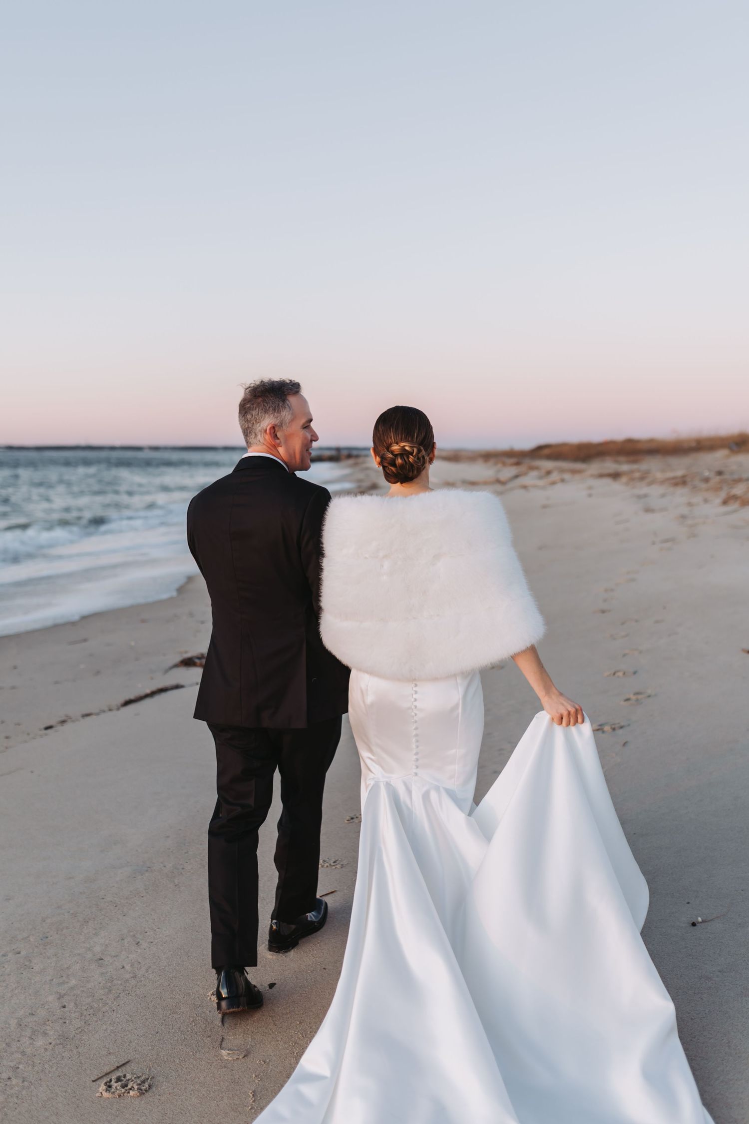 Bride and groom walking hand in hand on a beach at sunset, bride wearing white gown with fur wrap.