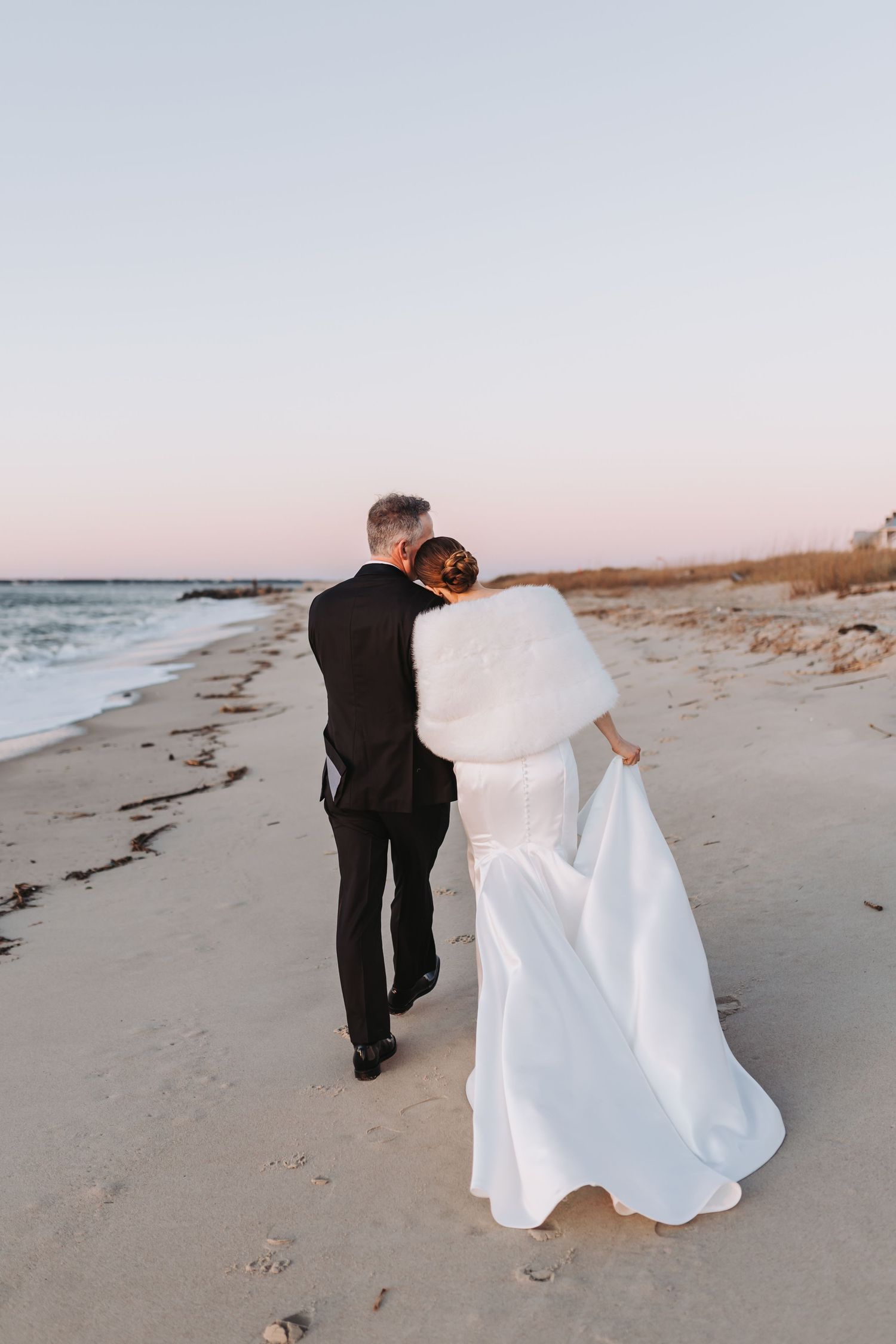 Bride and groom walk along beach at sunset, bride holding white gown train.