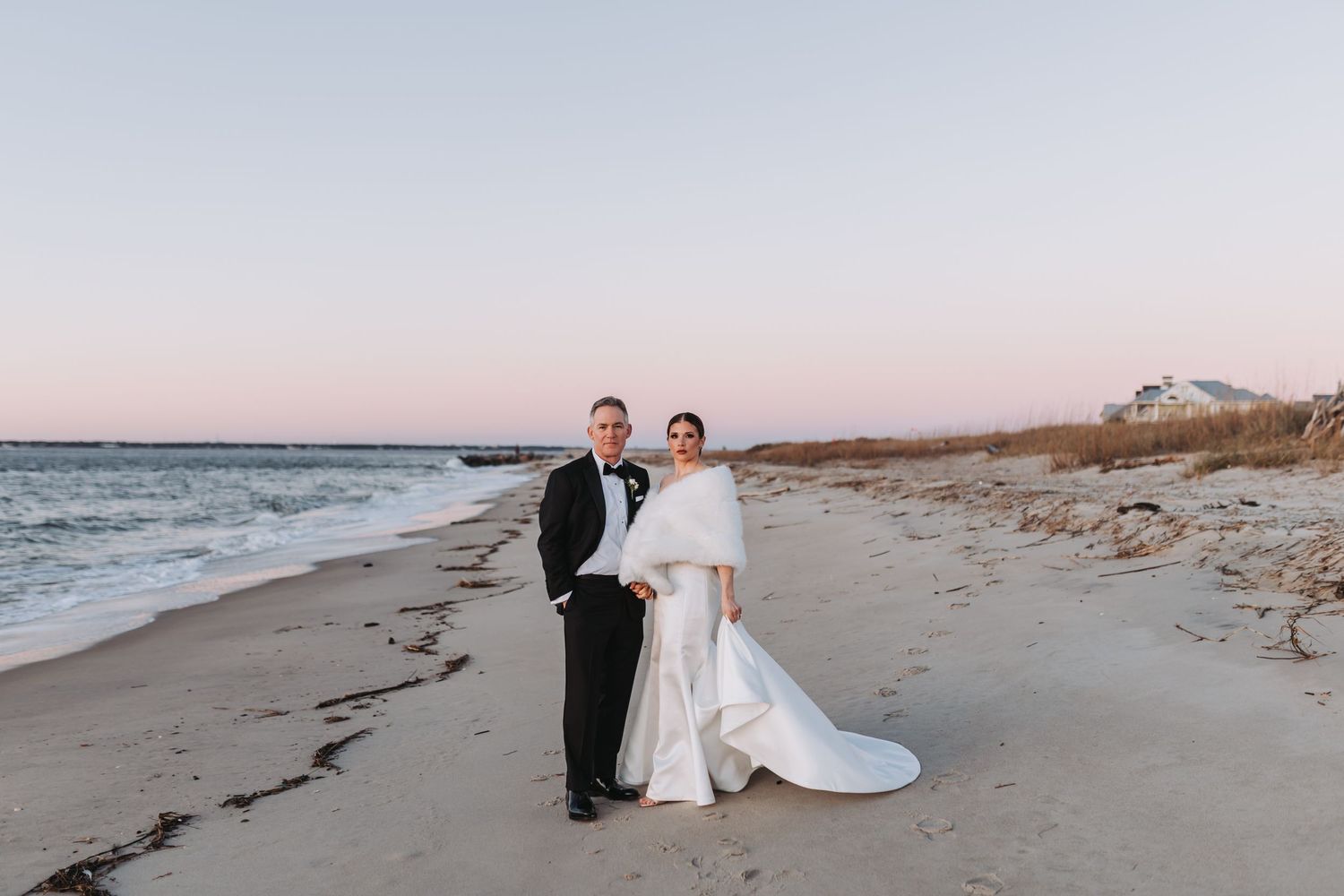 Bride and groom pose on a sandy beach at sunset, bride wearing a white gown with fur wrap and long train.