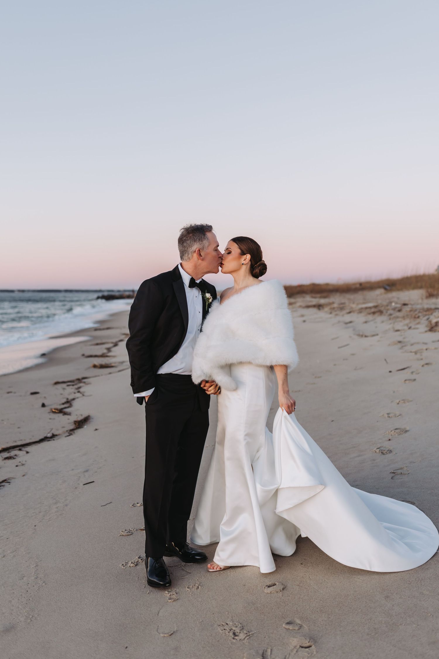 Bride and groom share a kiss on a sandy beach at sunset, she in a white gown with fur wrap, he in a black tuxedo.