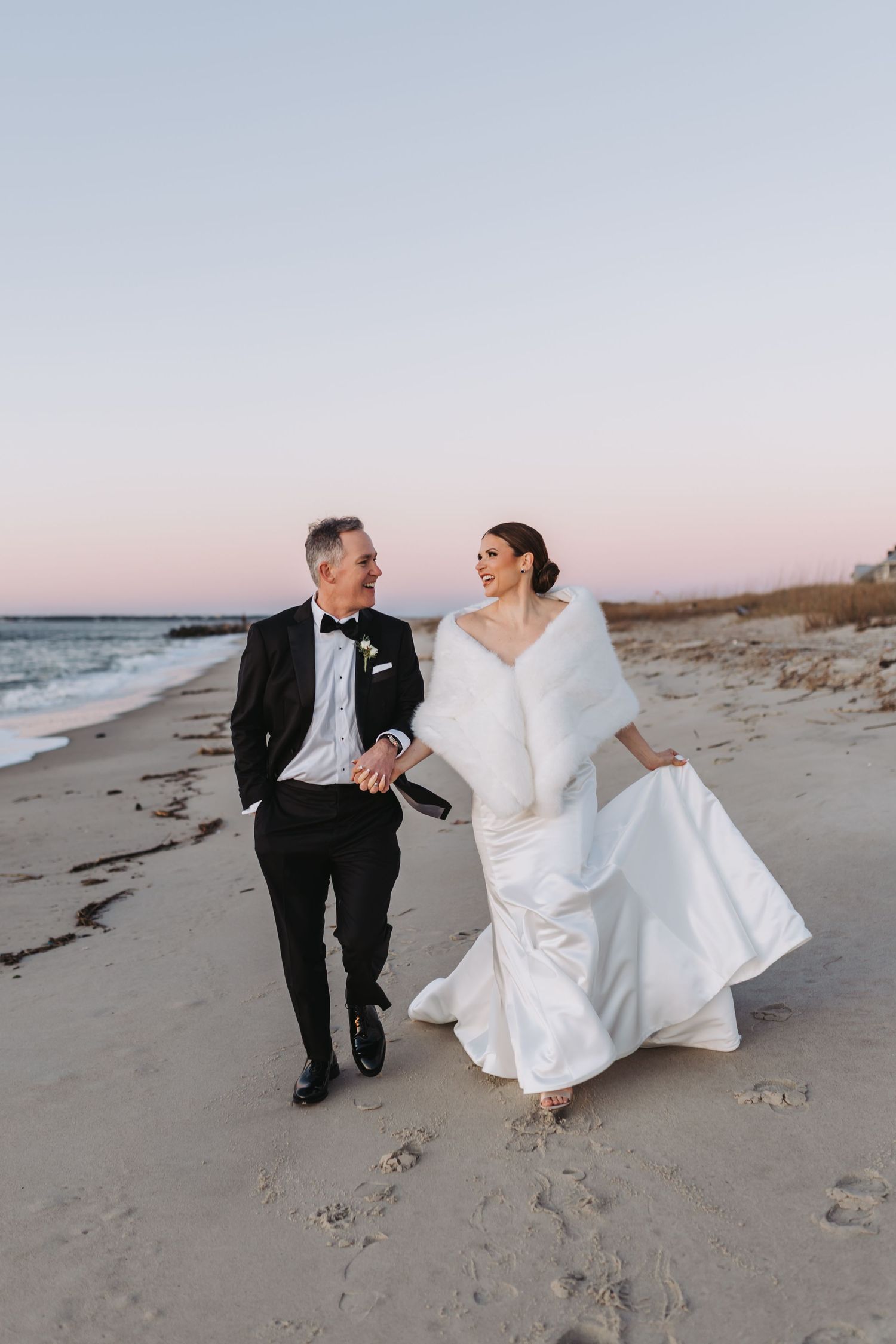 Bride and groom walking hand in hand on a beach at sunset, bride in white gown with fur wrap.