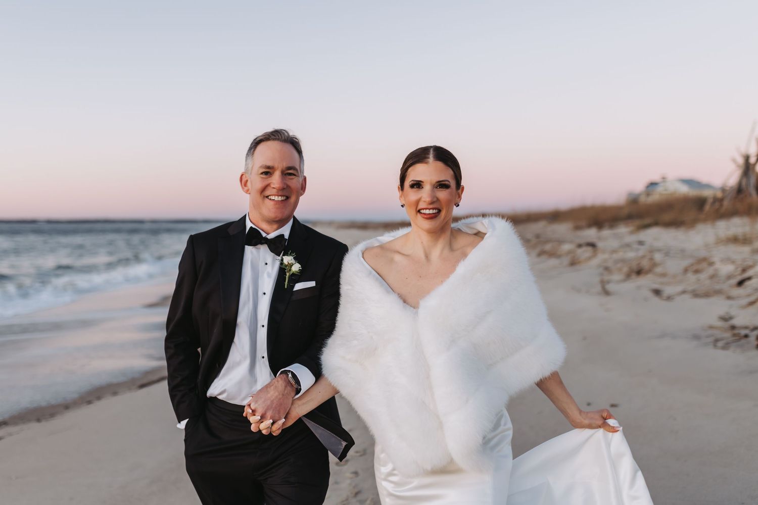 Bride and groom smiling on a beach at sunset, she in a white gown with fur wrap, he in a black tuxedo.