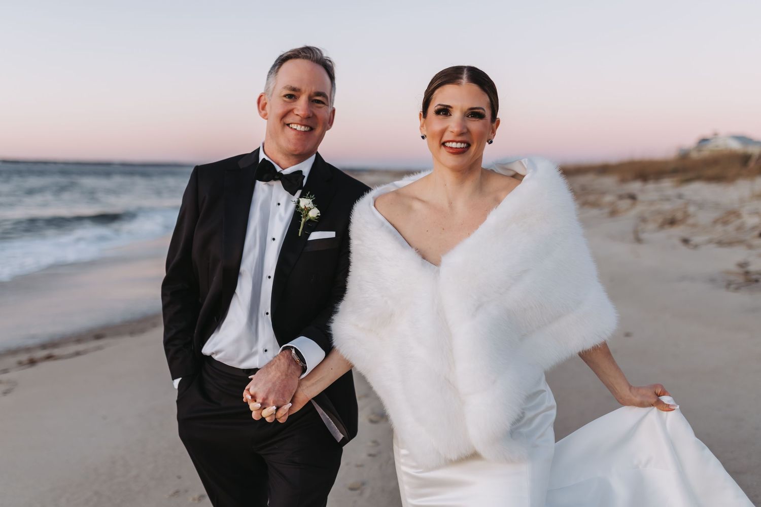 Couple in wedding attire smiling on a beach at sunset, groom in tuxedo and bride in white gown with fur wrap.