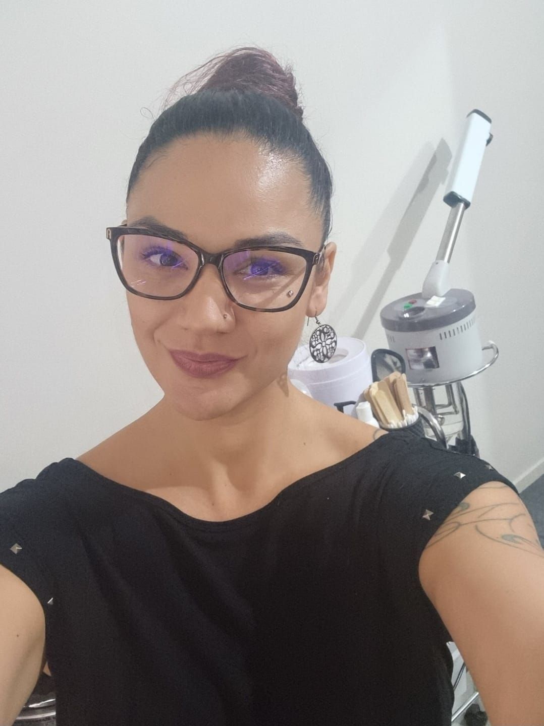 Woman with glasses and dark hair in bun wearing black top, taking selfie in beauty salon with laser equipment.