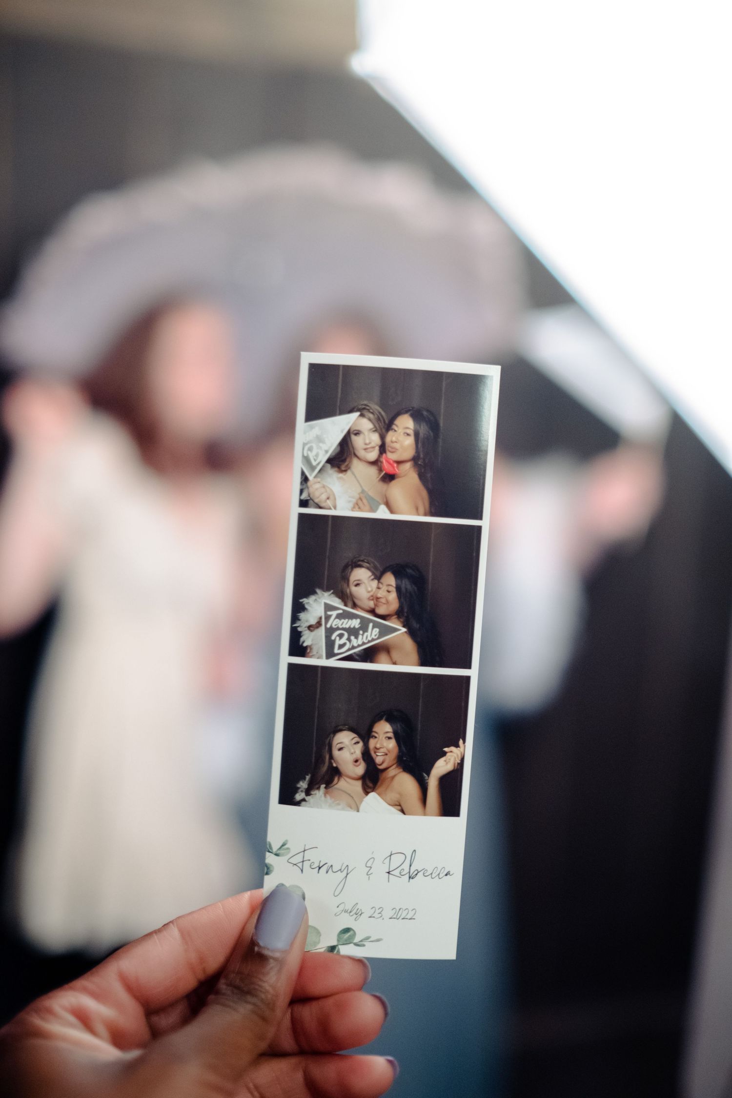 Hand holding a photo booth strip from a wedding, showing two women posing together, with bride details visible.