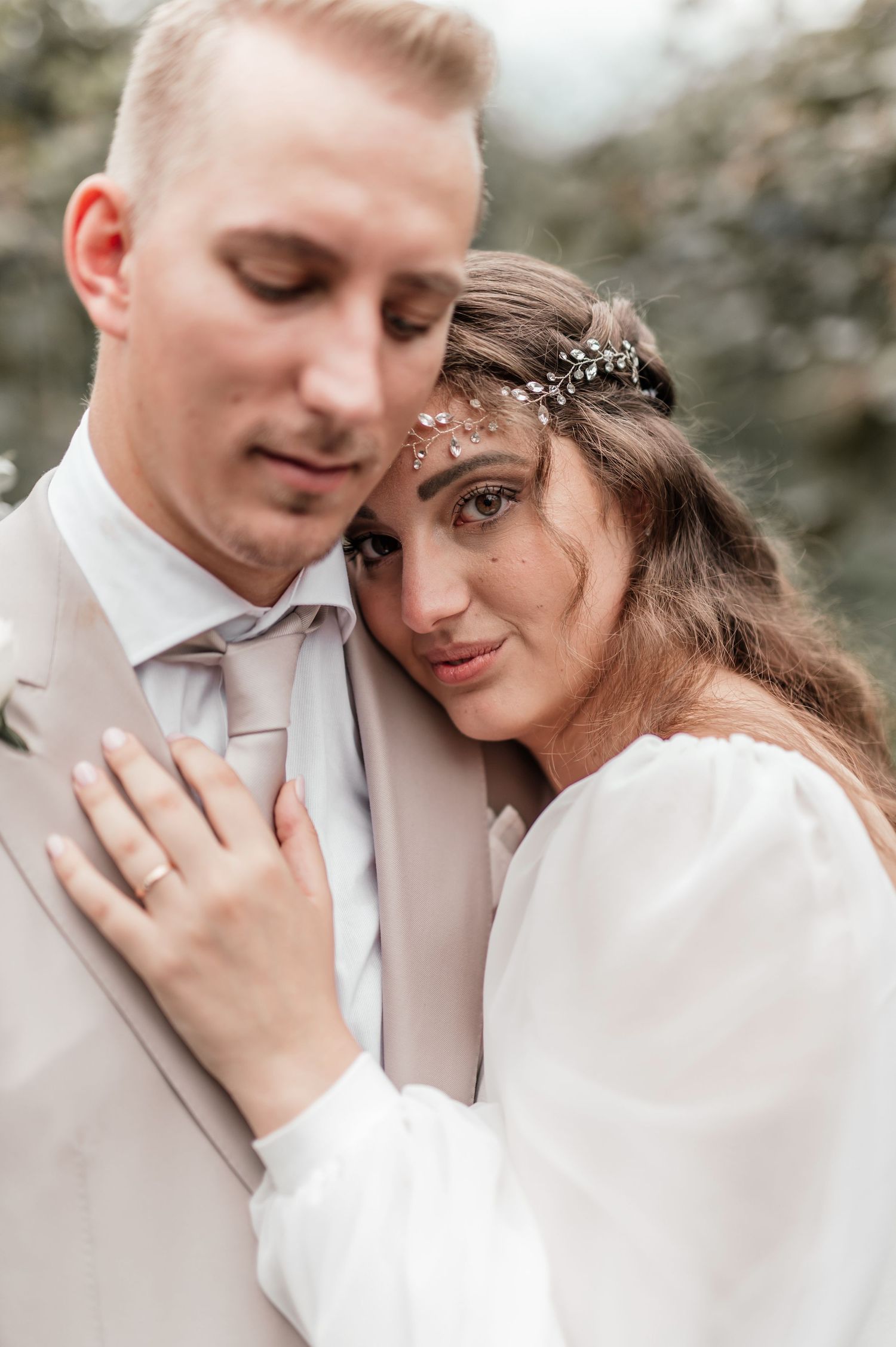 Intimate wedding portrait of bride in white dress leaning on groom in beige suit outdoors.