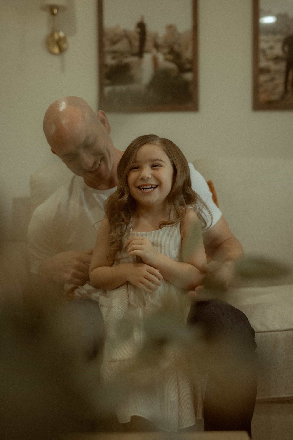 Bald father laughing and hugging his joyful young daughter on a couch in a cozy home.