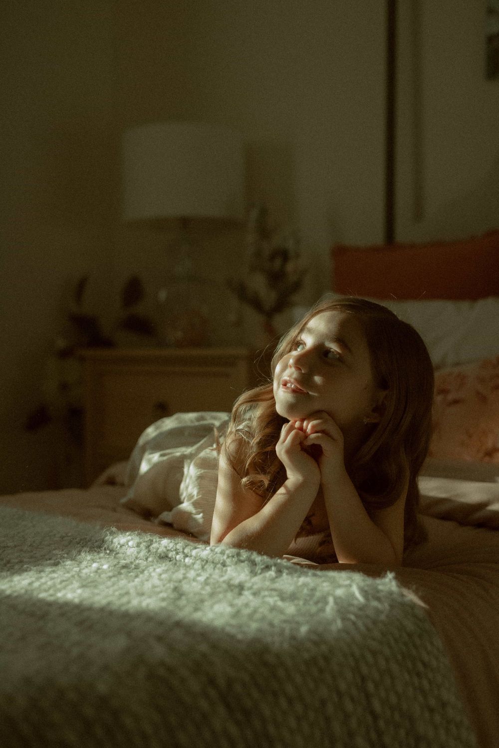 Young girl lying on a bed gazing upward with a gentle smile in warm bedroom lighting.