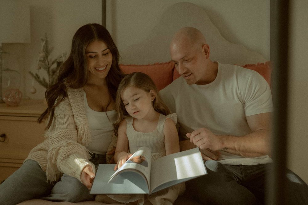 Family of three sitting together reading a book in a cozy bedroom setting.