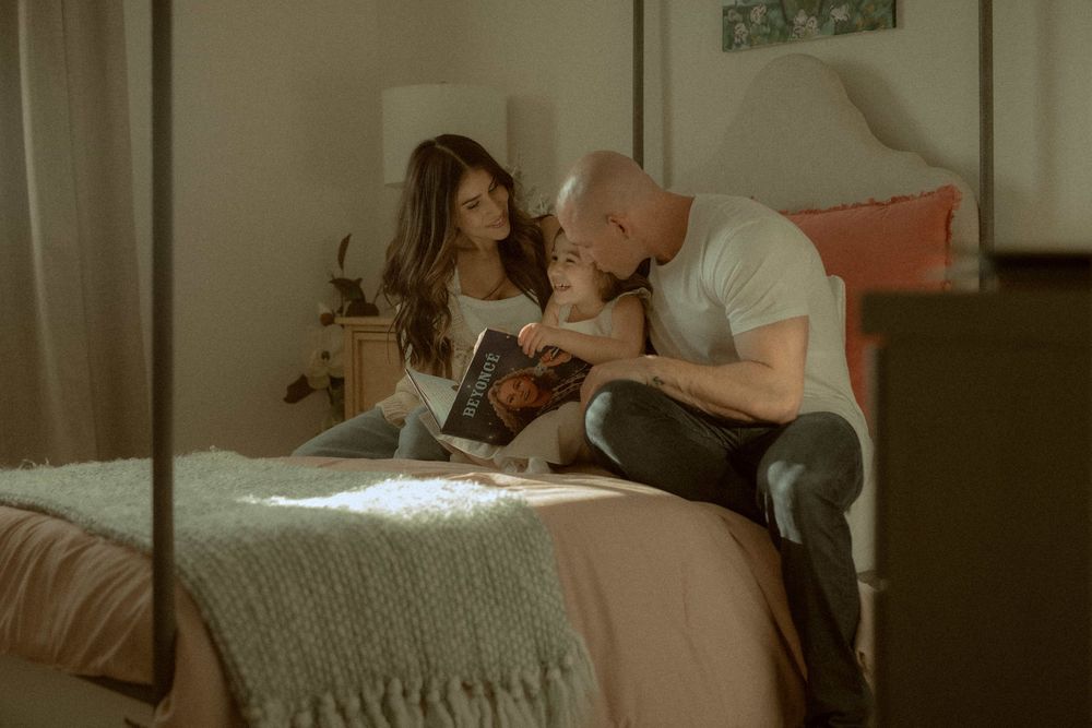 Mother and father sit on a bed reading a book together in a softly lit bedroom.