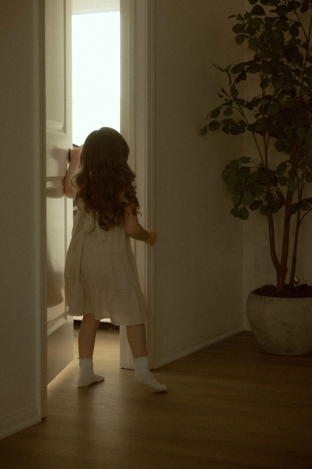 Young girl in white dress and socks opening a bright door in a softly lit hallway with a potted plant nearby.