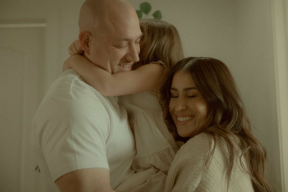 Bald man and dark-haired woman smiling warmly as a child hugs them both in a bright cozy room.