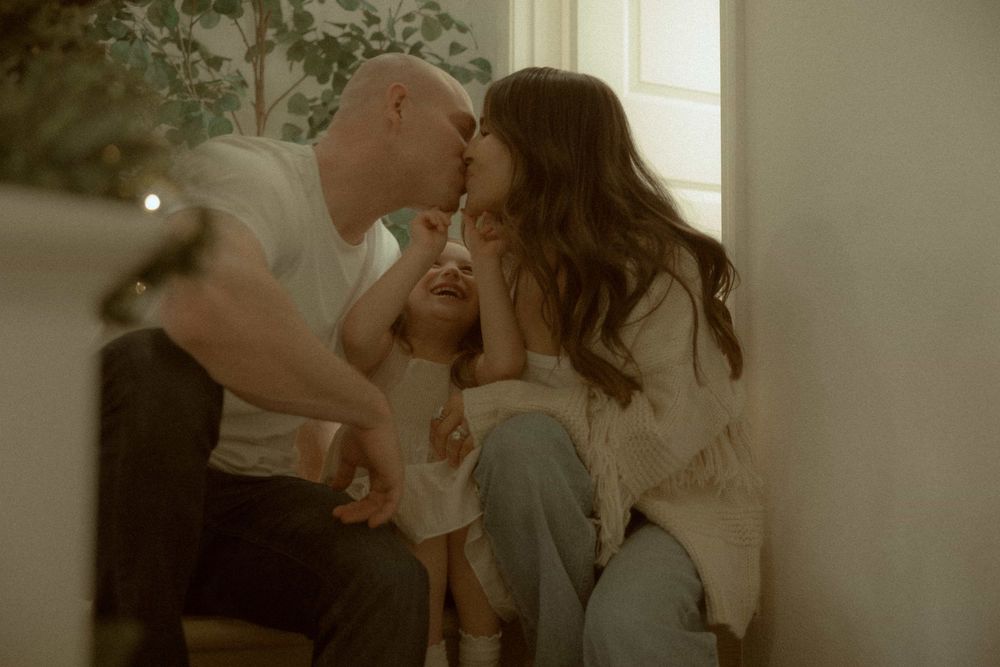 Parents kissing their young child on the cheeks simultaneously while sitting together near a bright window.