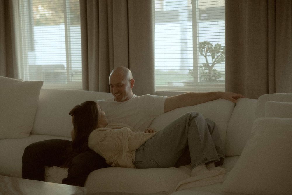 Man and woman relaxing together on a white sofa in a bright living room with large windows.