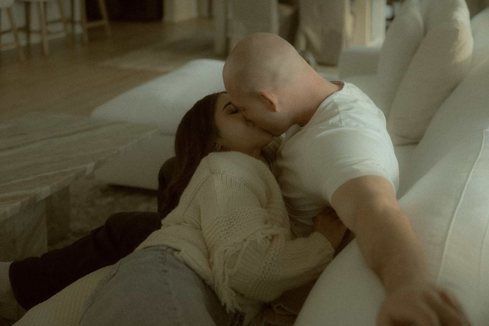 Couple sharing a romantic kiss while lying together on a white couch in a warmly lit room.