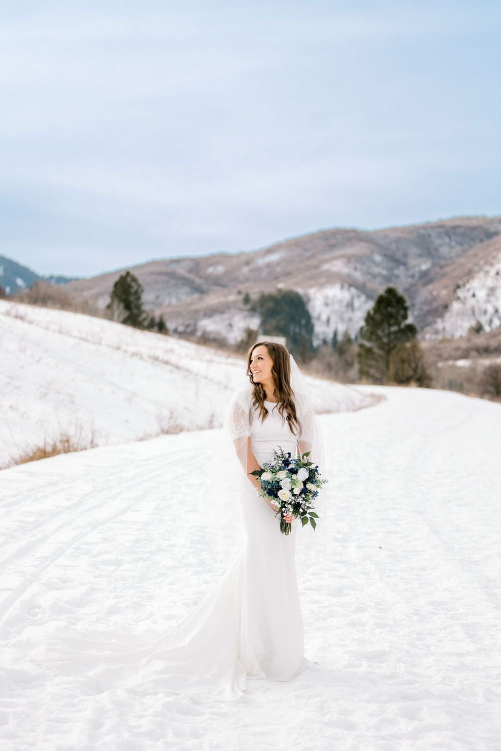 Bride in white gown holding bouquet stands on snowy mountain road in winter wedding portrait.