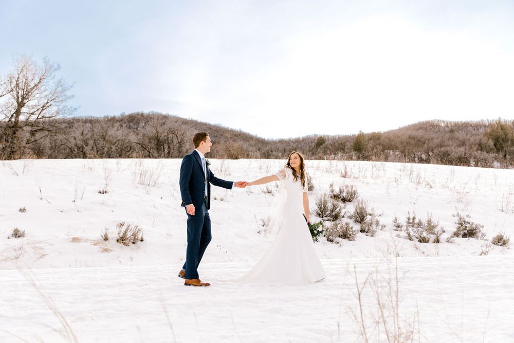 Bride and groom hold hands in snowy winter landscape with mountains, bride in flowing white gown, groom in navy suit.